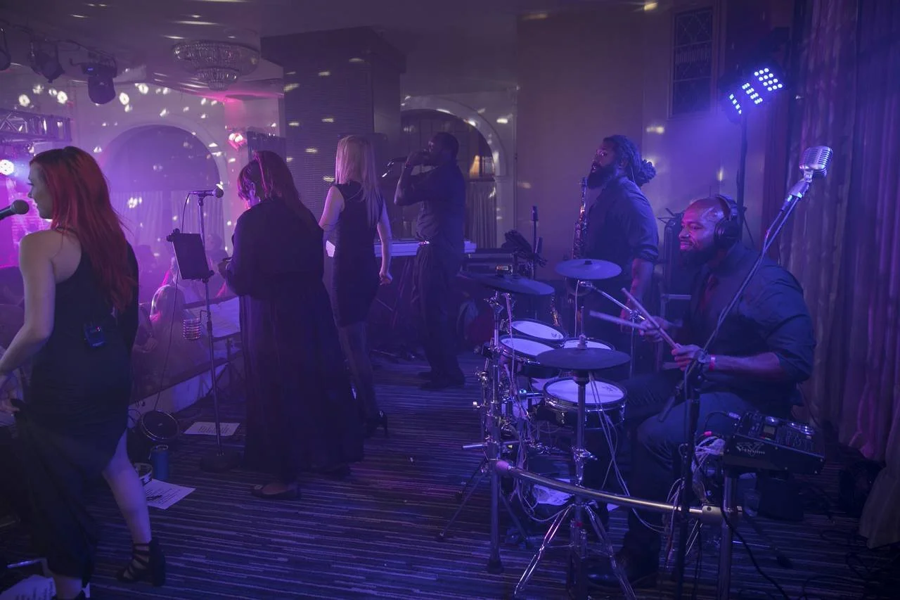 Live band performing on stage in dim lighting with multiple singers and musicians playing instruments; drummer in foreground.