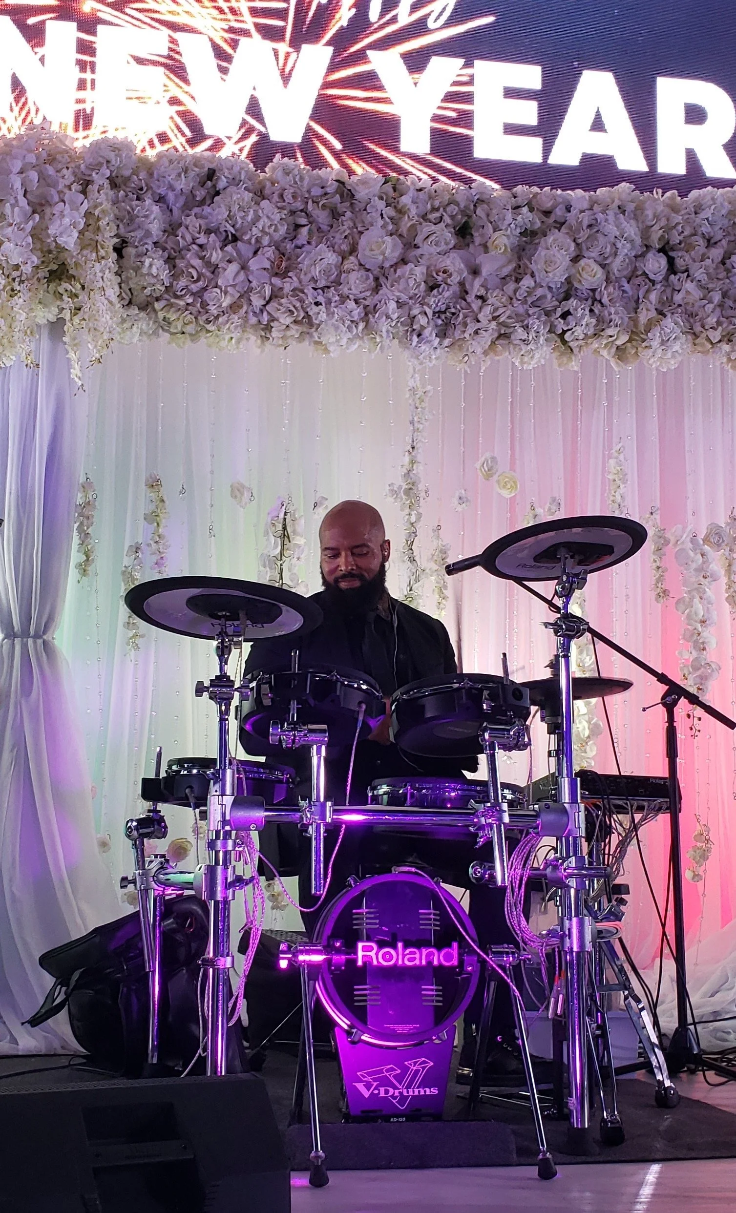 A drummer playing electronic drums, with a backdrop of white flowers and curtains, at a New Year's celebration event.