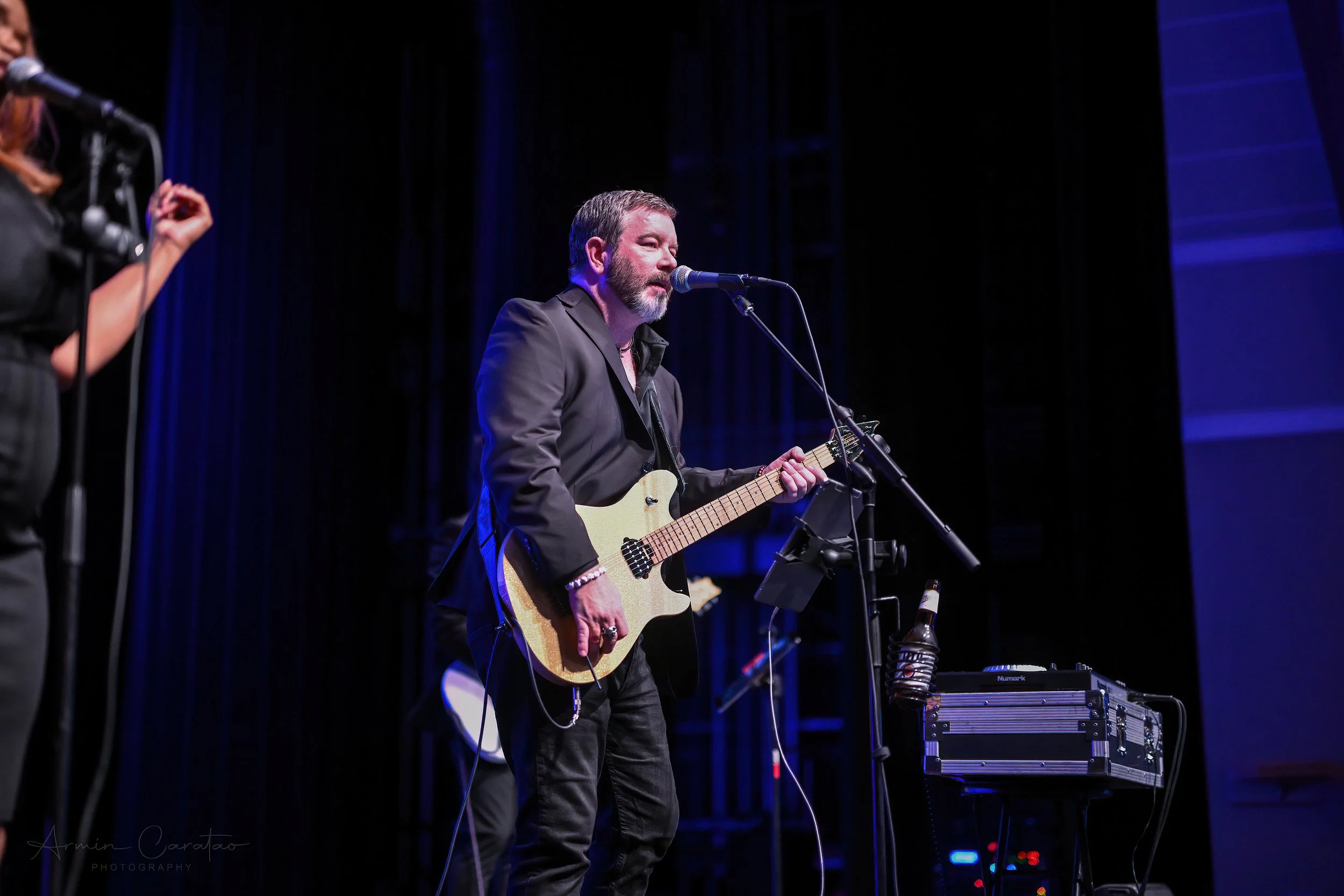 Musician in a black suit playing an electric guitar and singing into a microphone on stage.