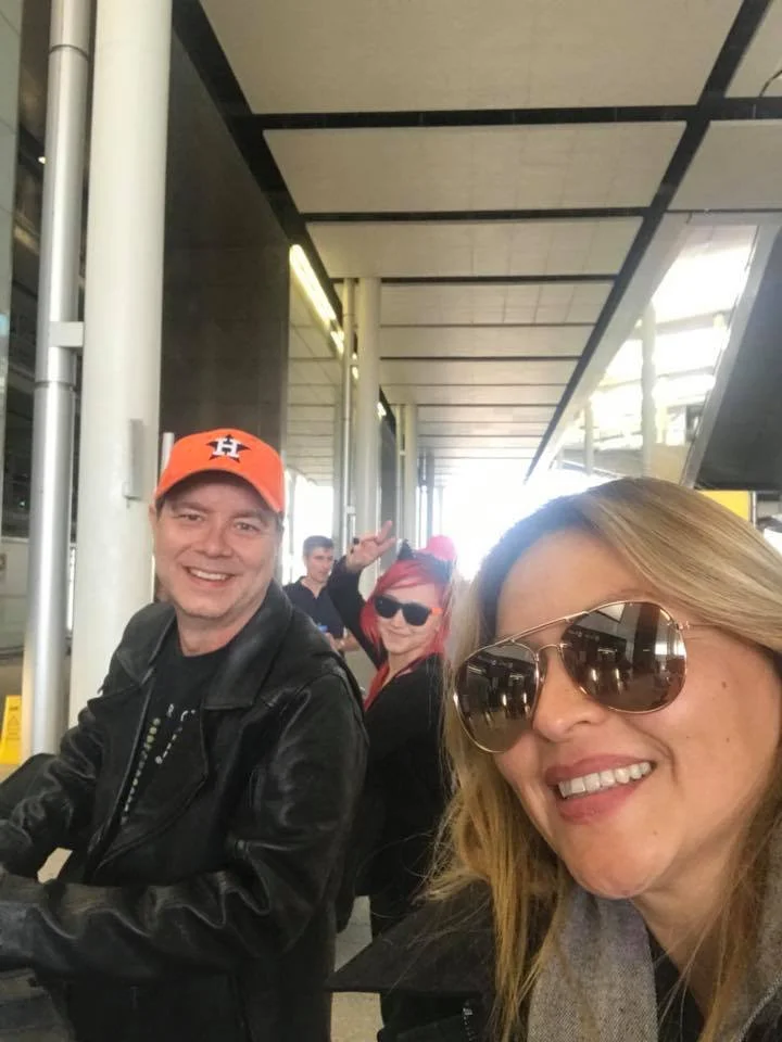 Group of people smiling at an airport, one wearing an orange sports cap, another with sunglasses giving a peace sign.