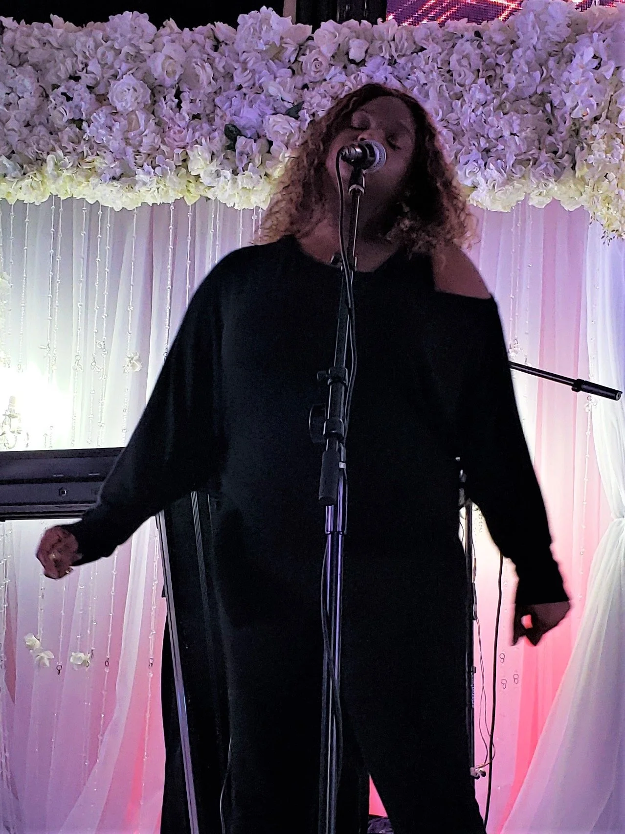 Woman singing on stage with floral backdrop and microphone.