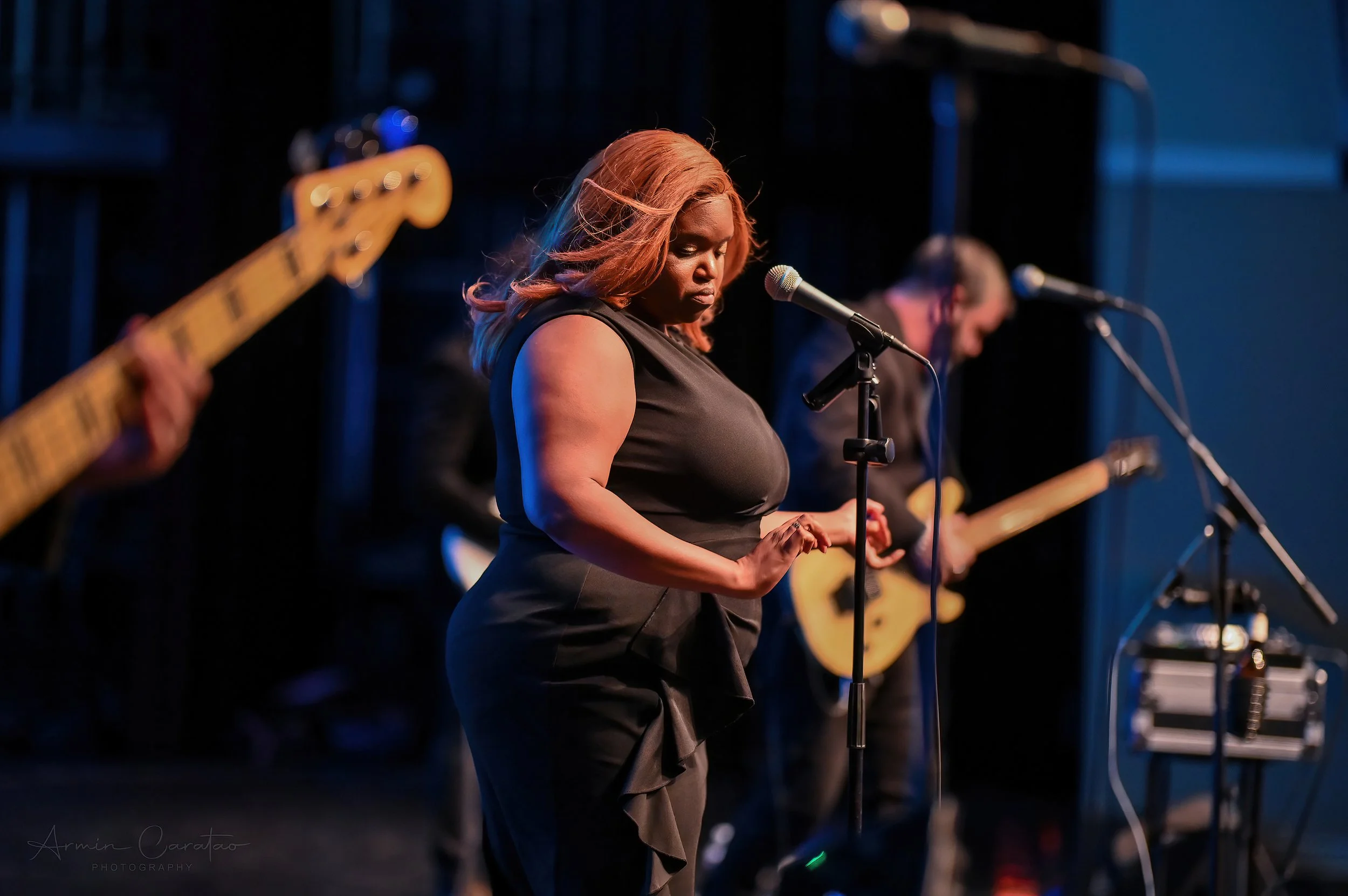 Woman singing on stage with musicians playing guitars in the background.