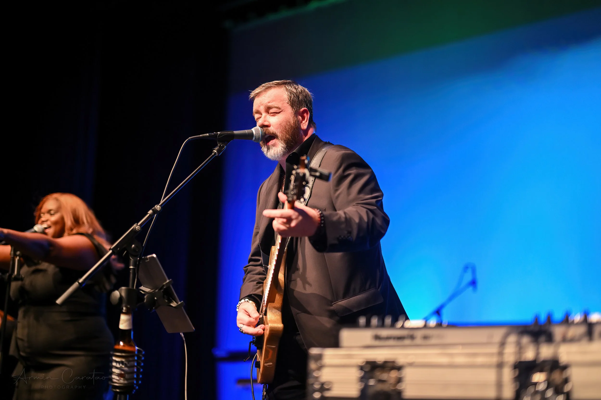 A male musician singing and playing guitar on stage with a female backup singer. The stage is lit with blue lighting, and audio equipment is visible in the foreground.