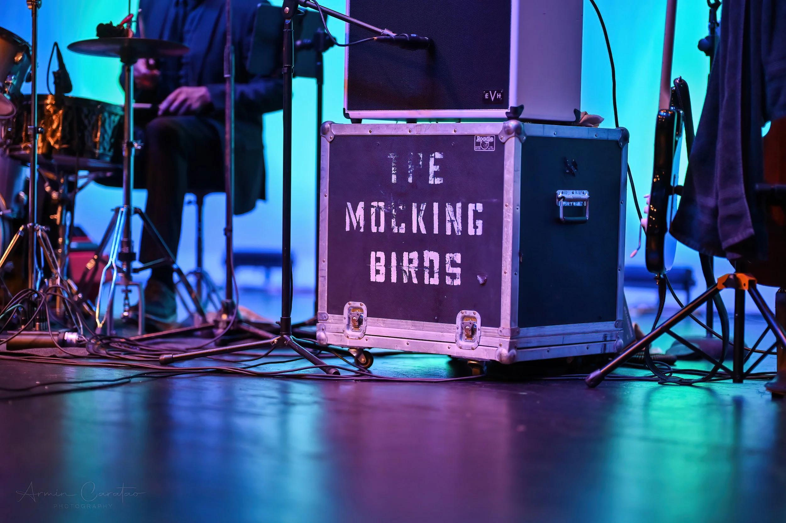 Music equipment and case labeled 'The Mocking Birds' on stage with drum set and speaker.