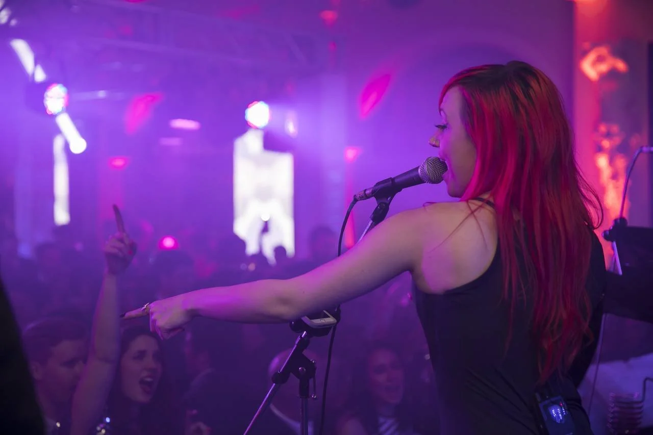 Singer with red hair performing on stage, microphone in hand, pointing at the audience in a vibrant, colorful nightclub setting.