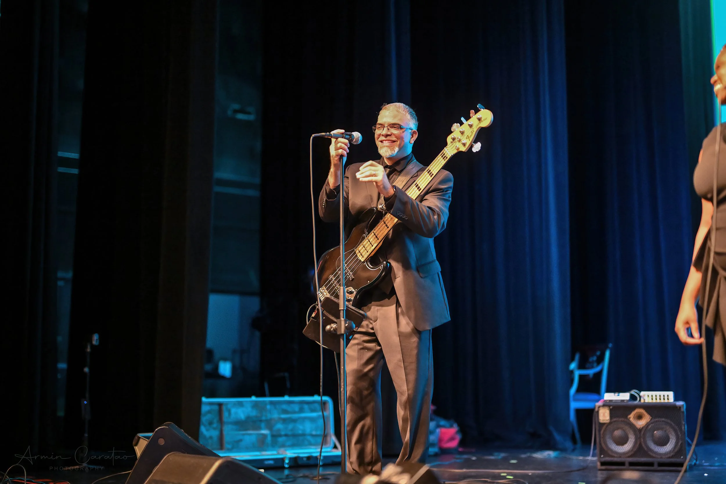 Man playing bass guitar on stage with microphone, wearing a suit, blue curtain background.