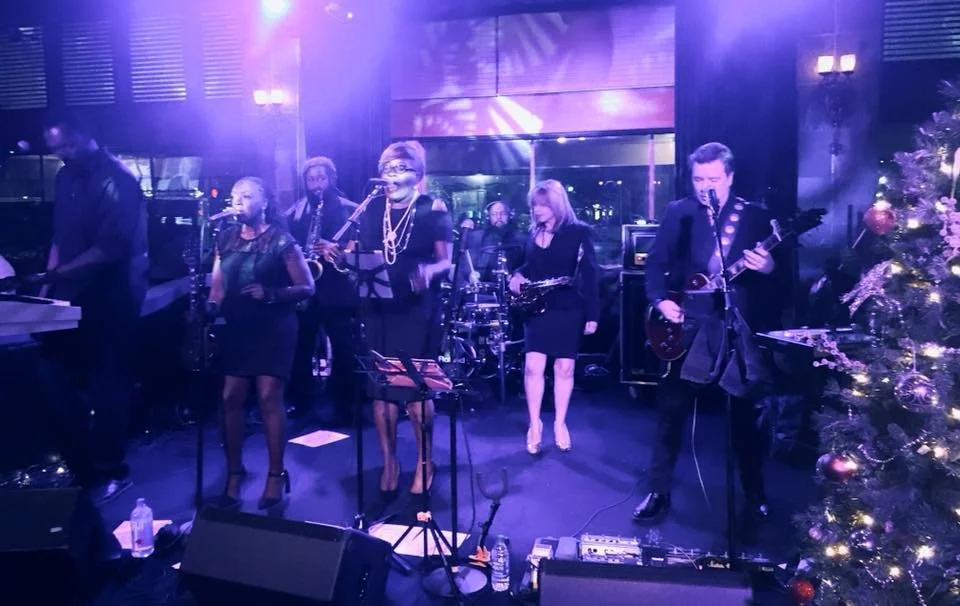A band performing live on stage with purple lighting, featuring vocalists, a guitarist, a keyboardist, and a saxophonist. A decorated Christmas tree is visible on the right side of the stage.