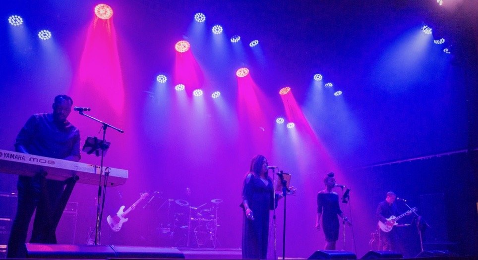 Band performing on stage with vibrant purple and blue lighting, featuring a keyboardist, two vocalists, a guitarist, and a drummer.