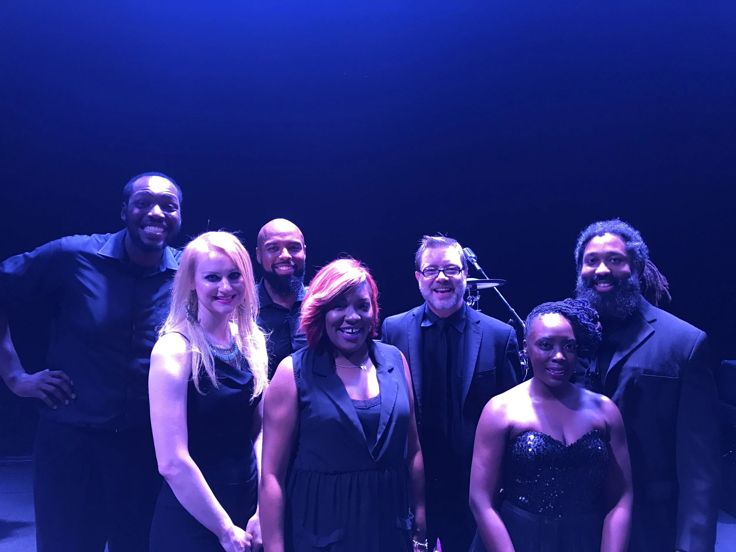 Group of seven people in black formal attire posing with musical equipment in background