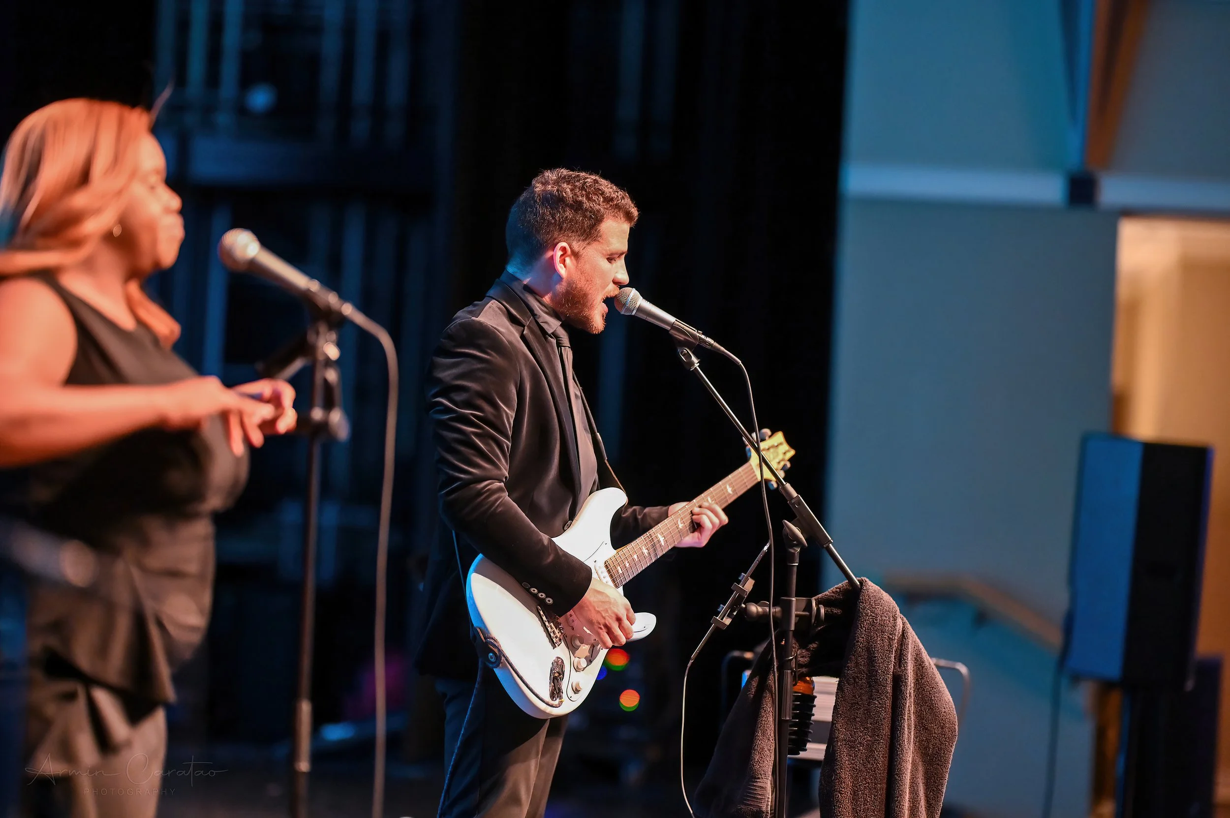 A musician in a suit playing an electric guitar and singing into a microphone on stage, accompanied by a backup singer.
