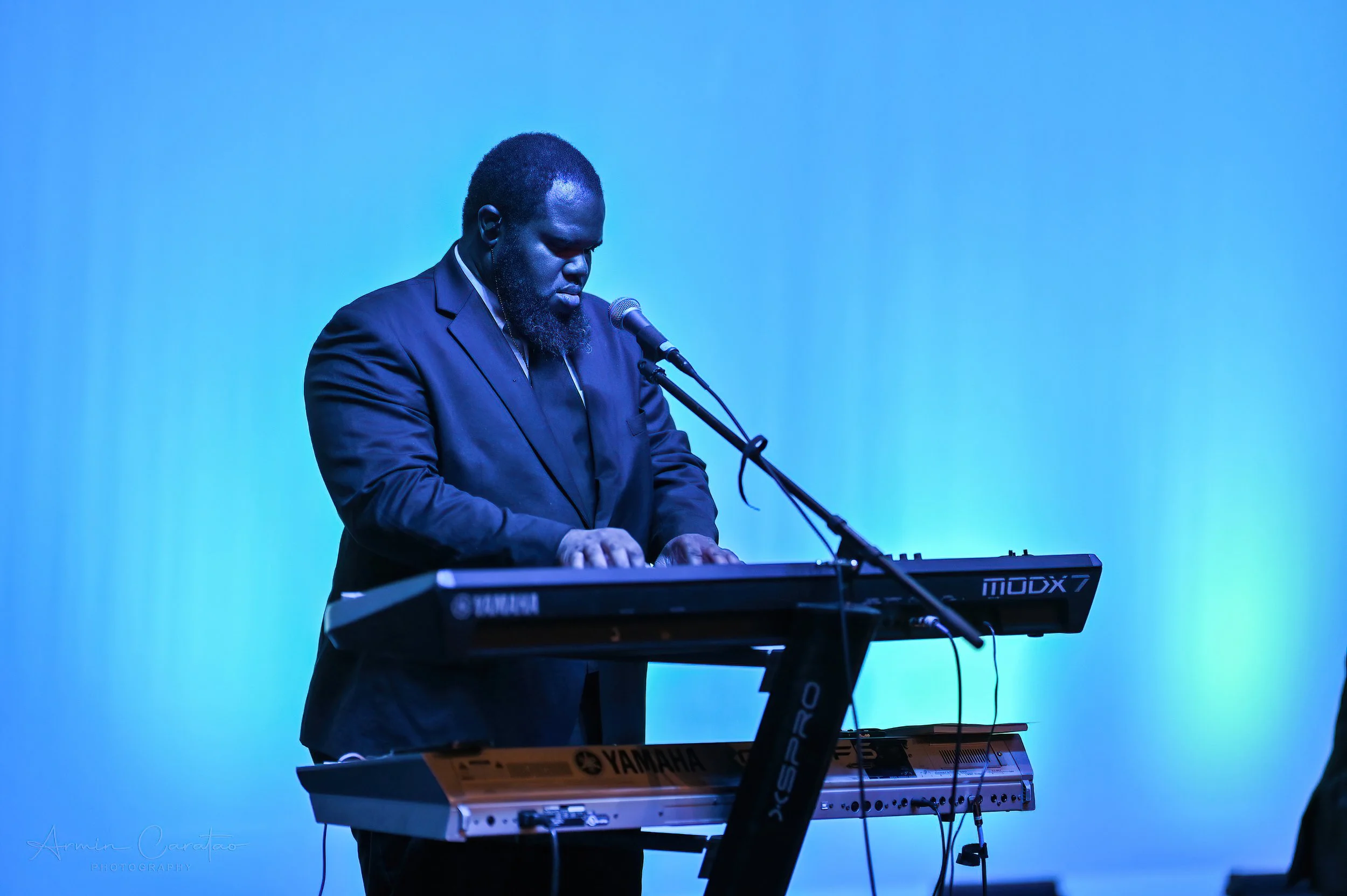 Musician in a suit playing a Yamaha keyboard on stage with blue lighting.