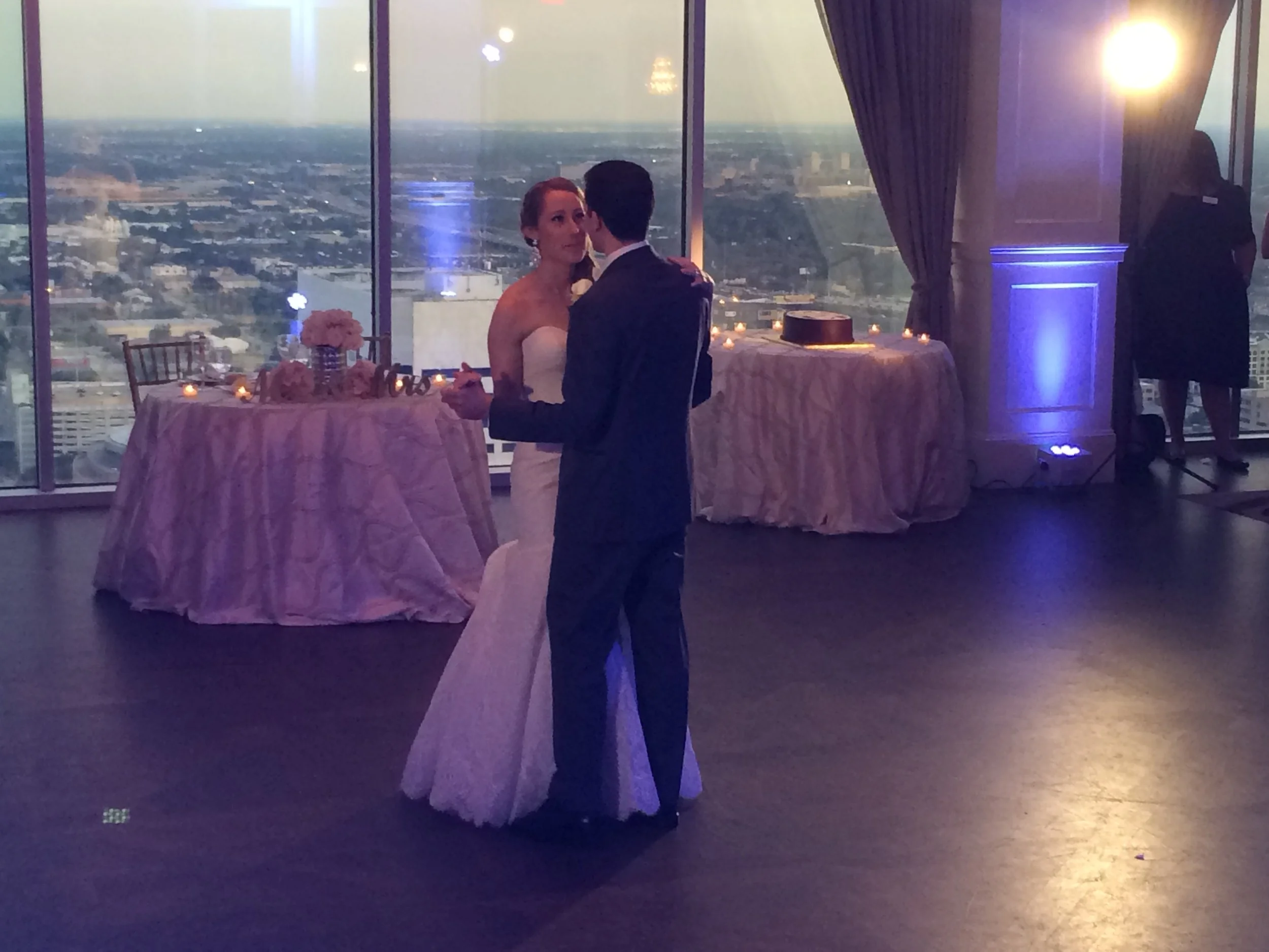 A couple dancing at a wedding reception in a venue with large windows, with a view of a cityscape at dusk. Tables decorated with flowers and candles are visible in the background.