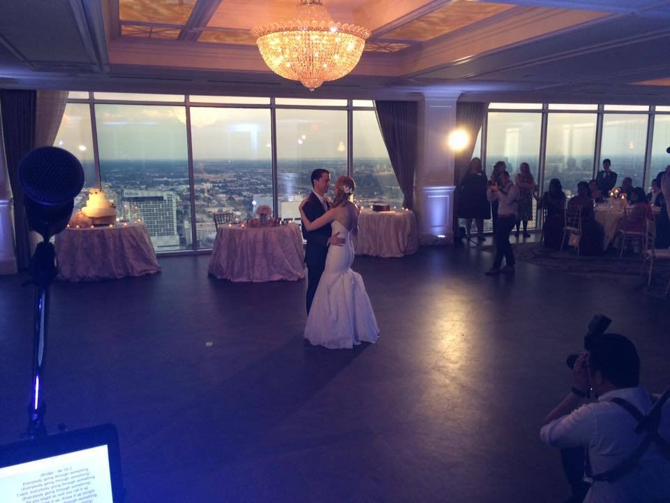 A couple is dancing in a large, elegant room with a city skyline view. The room has a chandelier, tables with guests, and a photographer capturing the moment.