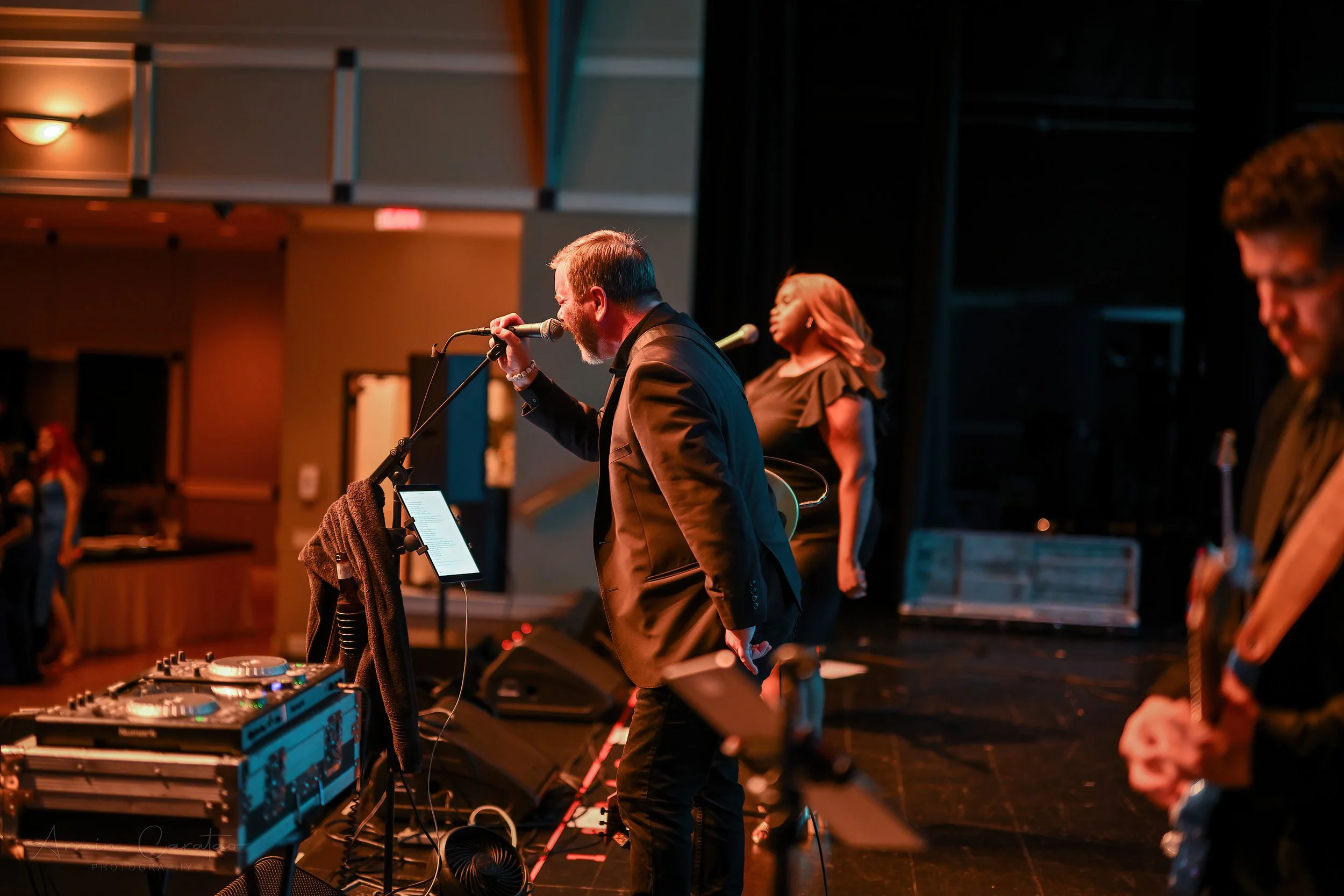 A band performing on stage with a male singer at the microphone and a female vocalist. A guitarist is visible in the foreground. There's audio equipment and stage lighting.