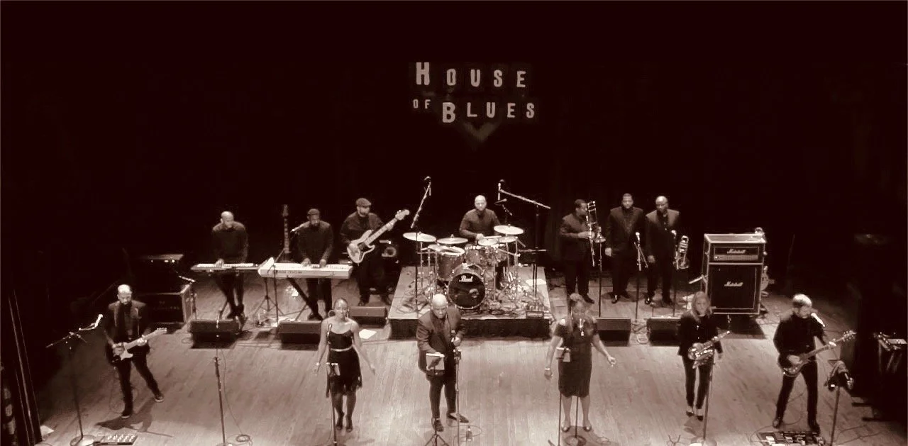 Band performing on stage at House of Blues, featuring musicians with guitars, keyboards, drums, and brass instruments.