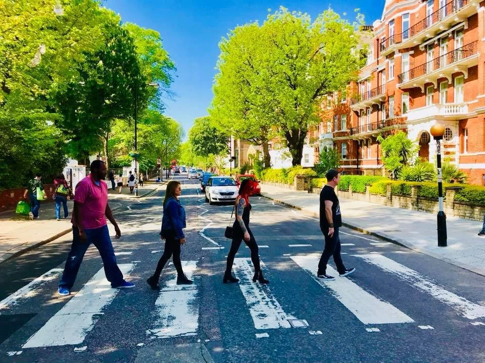 People walking on a zebra crossing on a sunny day, with buildings and trees in the background.