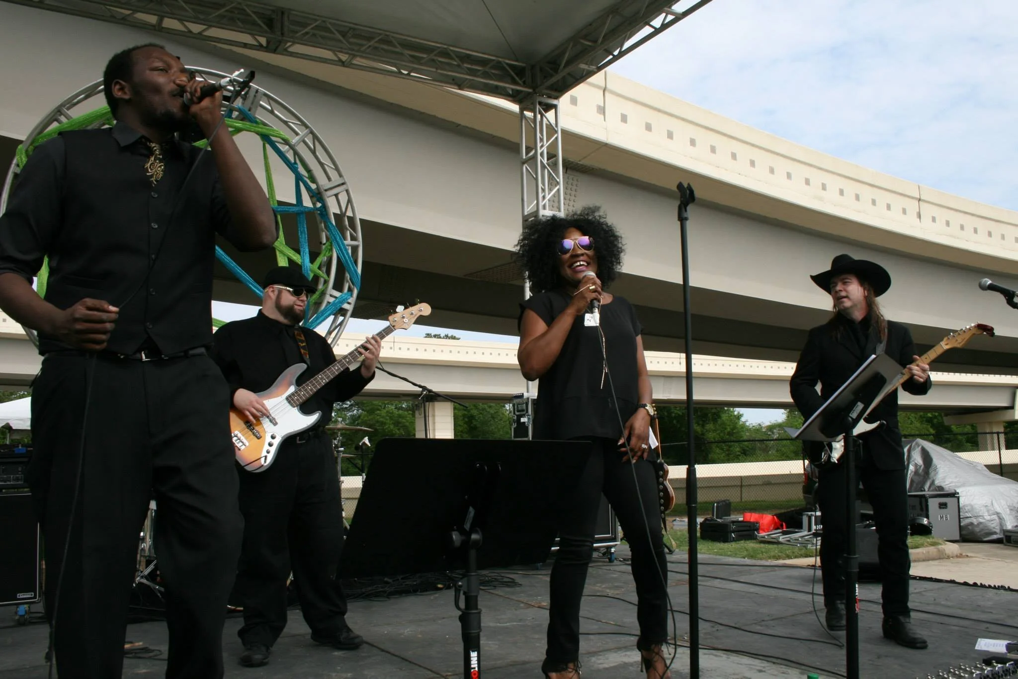 Band performing on outdoor stage, featuring male and female vocalists, one with a hat and playing guitar, another with sunglasses; bass player in background.
