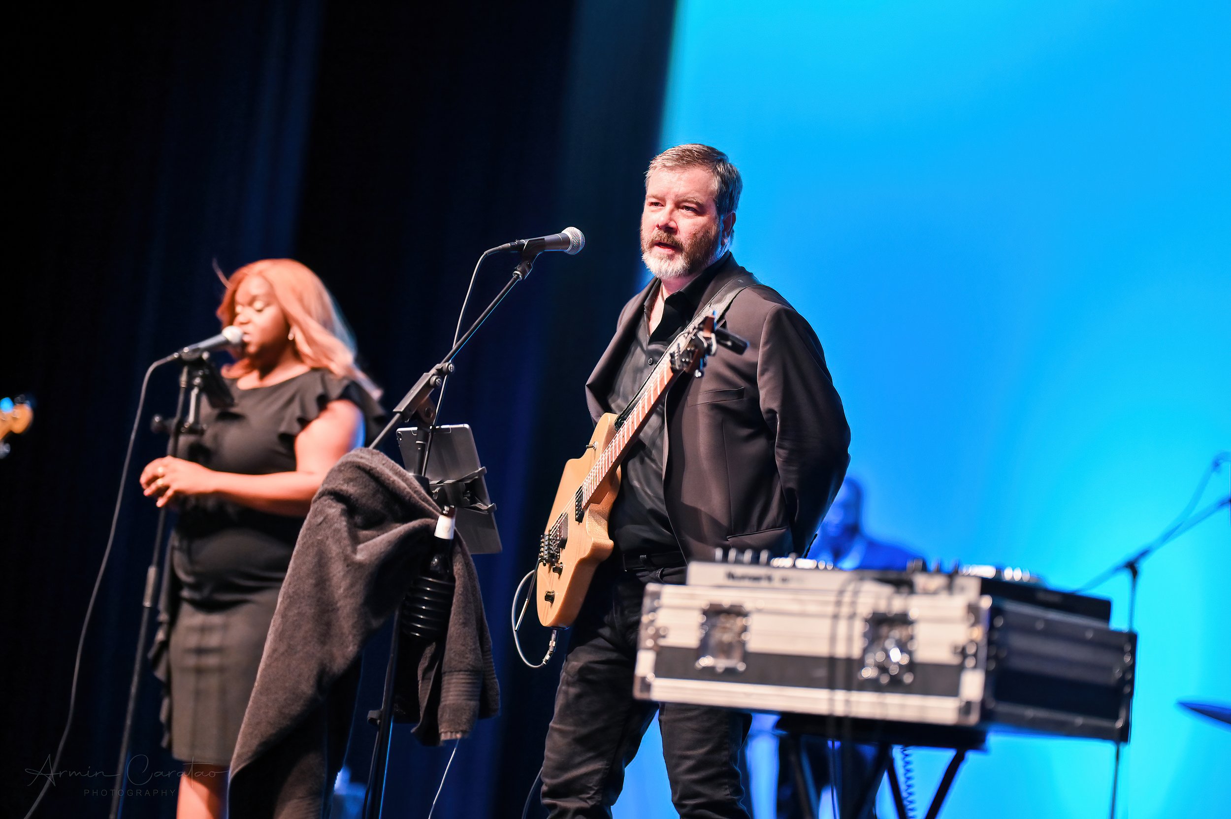 Musicians performing on stage, a woman singing into a microphone and a man playing guitar, with a blue background.