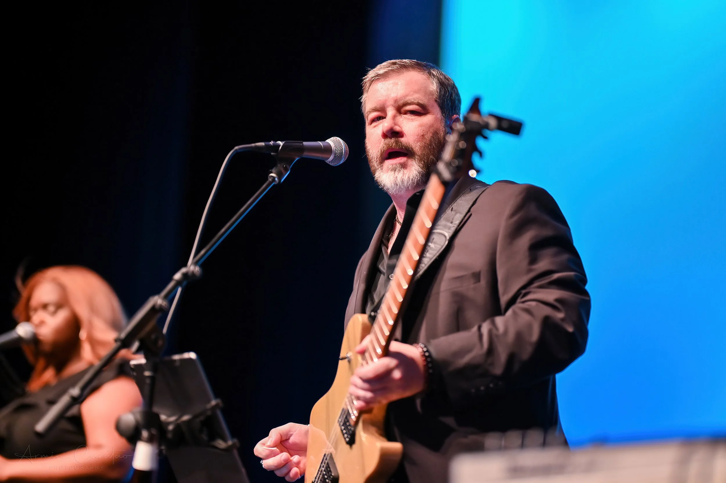 Man in a suit playing electric guitar on stage with a microphone, accompanied by a female singer in the background against a blue backdrop.