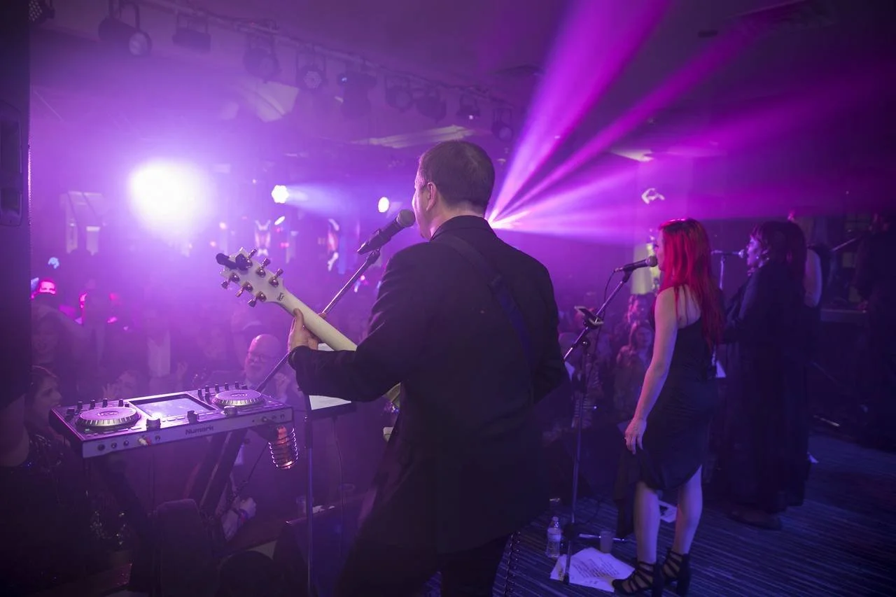 Live music performance with band on stage, colorful lighting, audience in background.