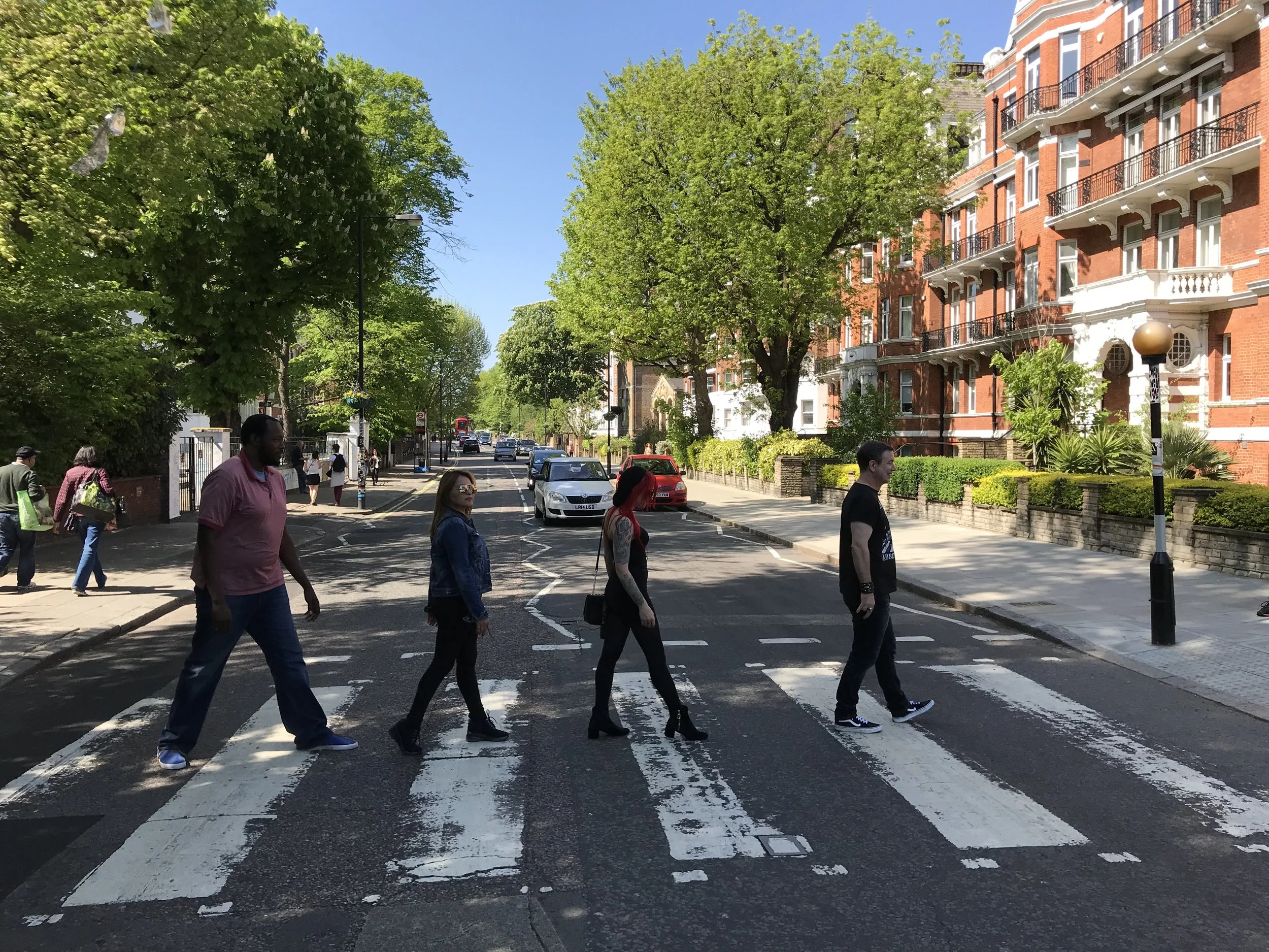 People walking across the iconic Abbey Road crosswalk in London on a sunny day, with red brick buildings and trees lining the street.