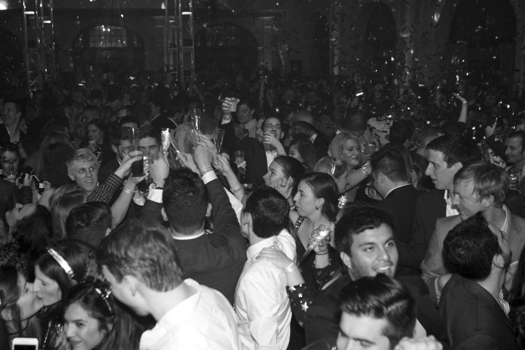 A crowded party with people celebrating, holding drinks, and cheering, in a black and white photo.