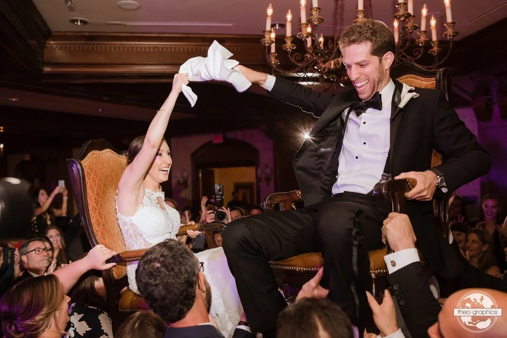 Bride and groom lifted on chairs during a wedding celebration, surrounded by guests, in a well-lit, elegant venue.
