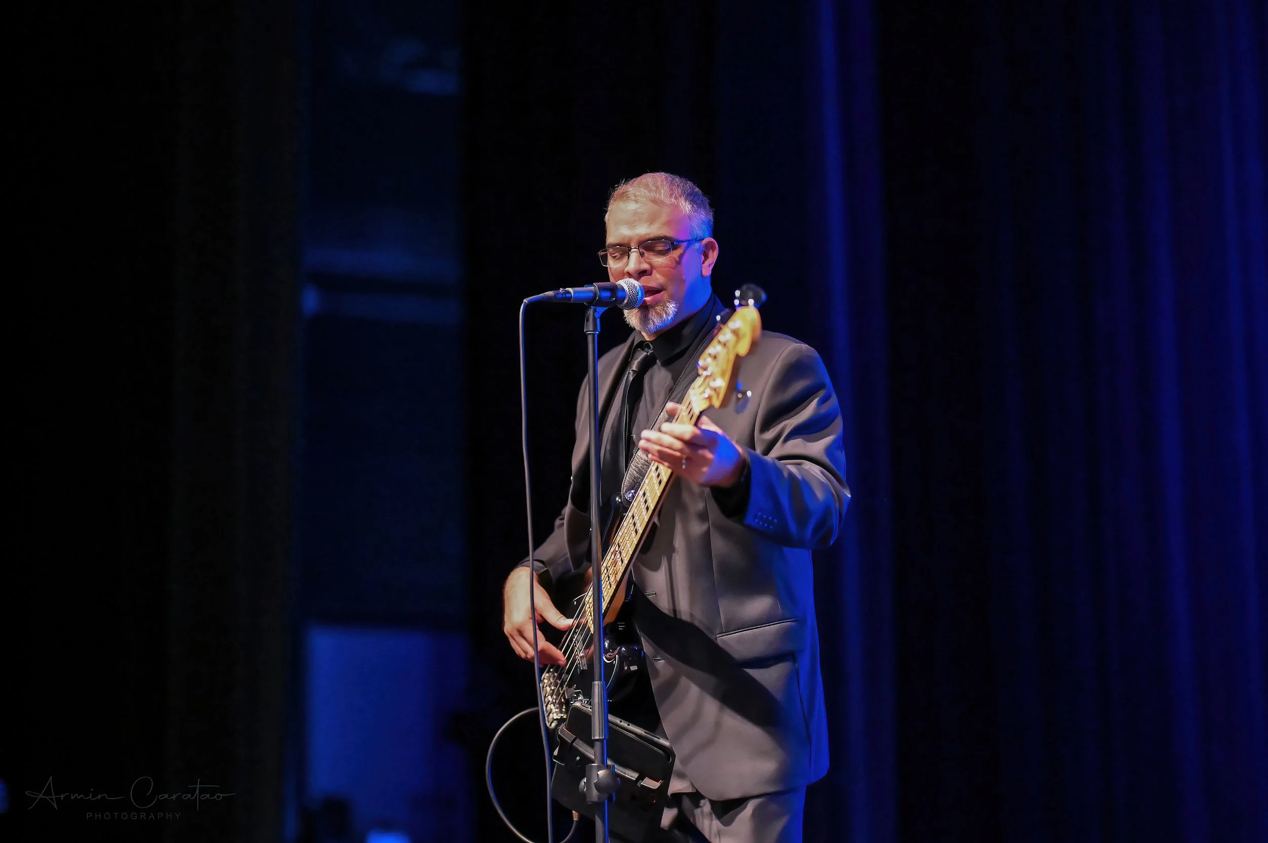 Musician in a suit playing a bass guitar on stage, singing into a microphone, against a dark curtain backdrop.