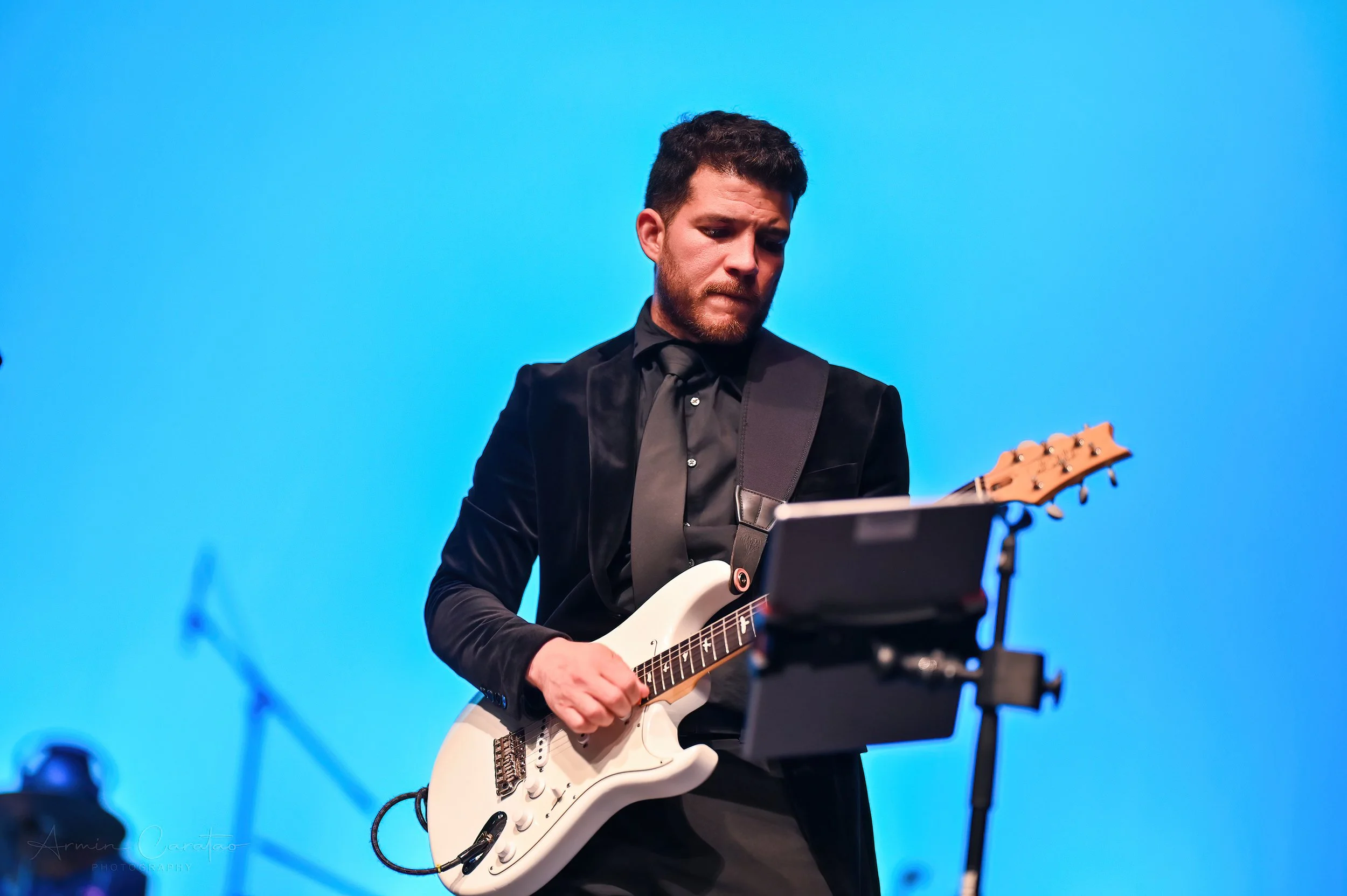 Man in a suit playing an electric guitar on stage with a blue background.