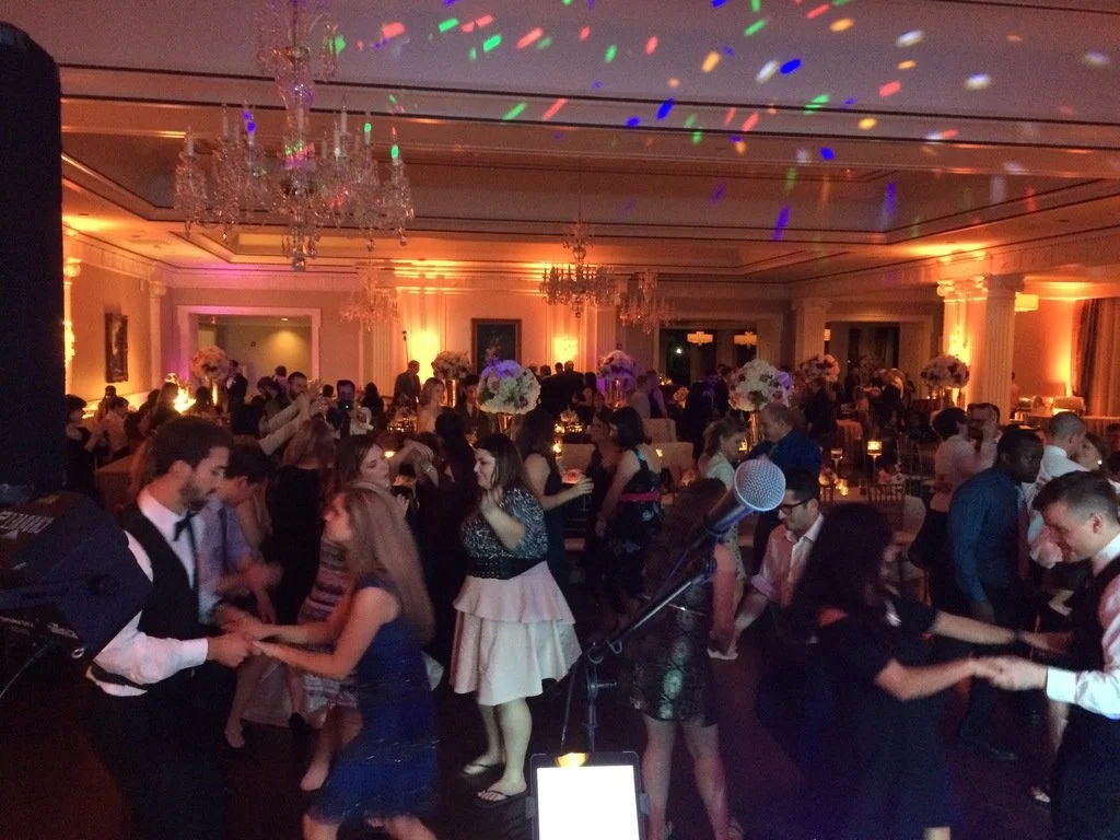 A lively indoor dance floor scene at an elegant event. People are dancing energetically in a large, well-lit room decorated with chandeliers and floral centerpieces. The atmosphere is festive with colorful light reflections on the ceiling.