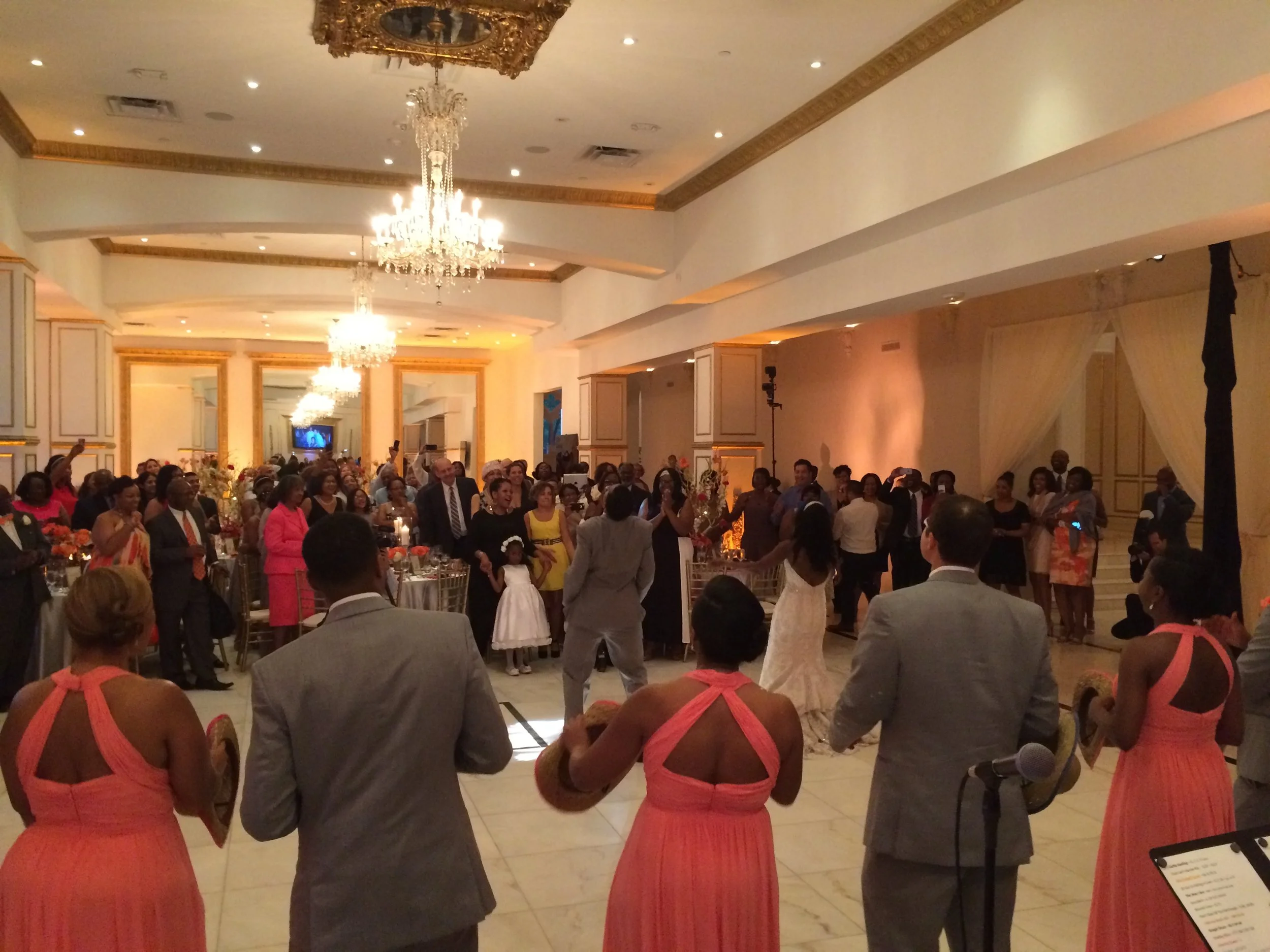 A crowded wedding reception in an elegant hall with chandeliers. Guests are gathered around a dancing couple in the center. The bridal party, dressed in pink and gray, is in the foreground, holding tambourines. The decor features gold accents and flo