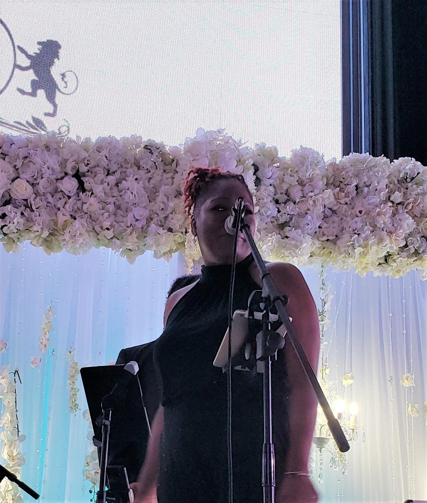 A woman in a black dress singing into a microphone, standing on a stage decorated with white flowers. A silhouette of a figure with a hoop is visible on a large screen behind her.