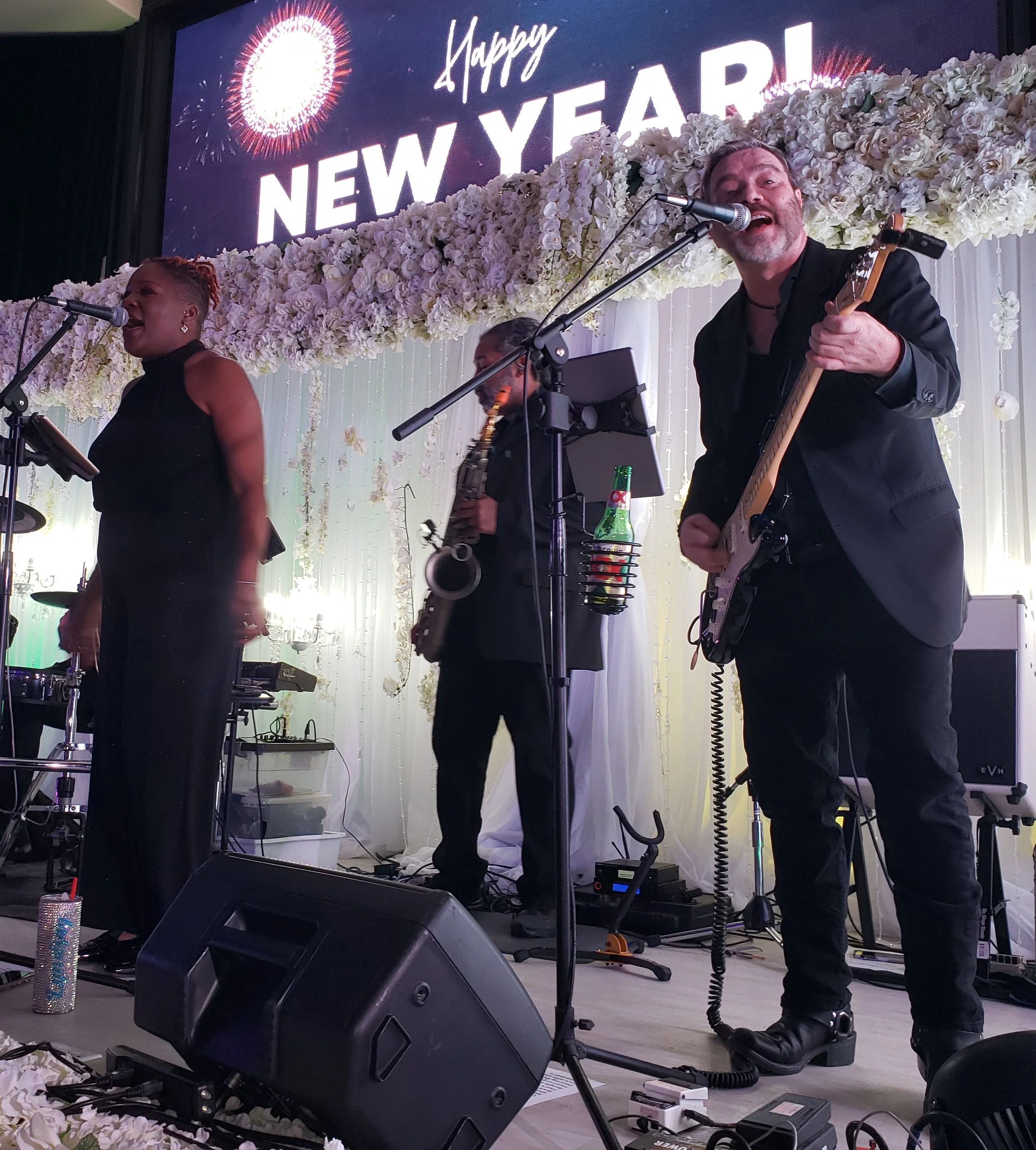 Band performing at a New Year's celebration, with a female vocalist, a saxophonist, and a male guitarist. The stage is decorated with white flowers and a "Happy New Year" sign with fireworks in the background.