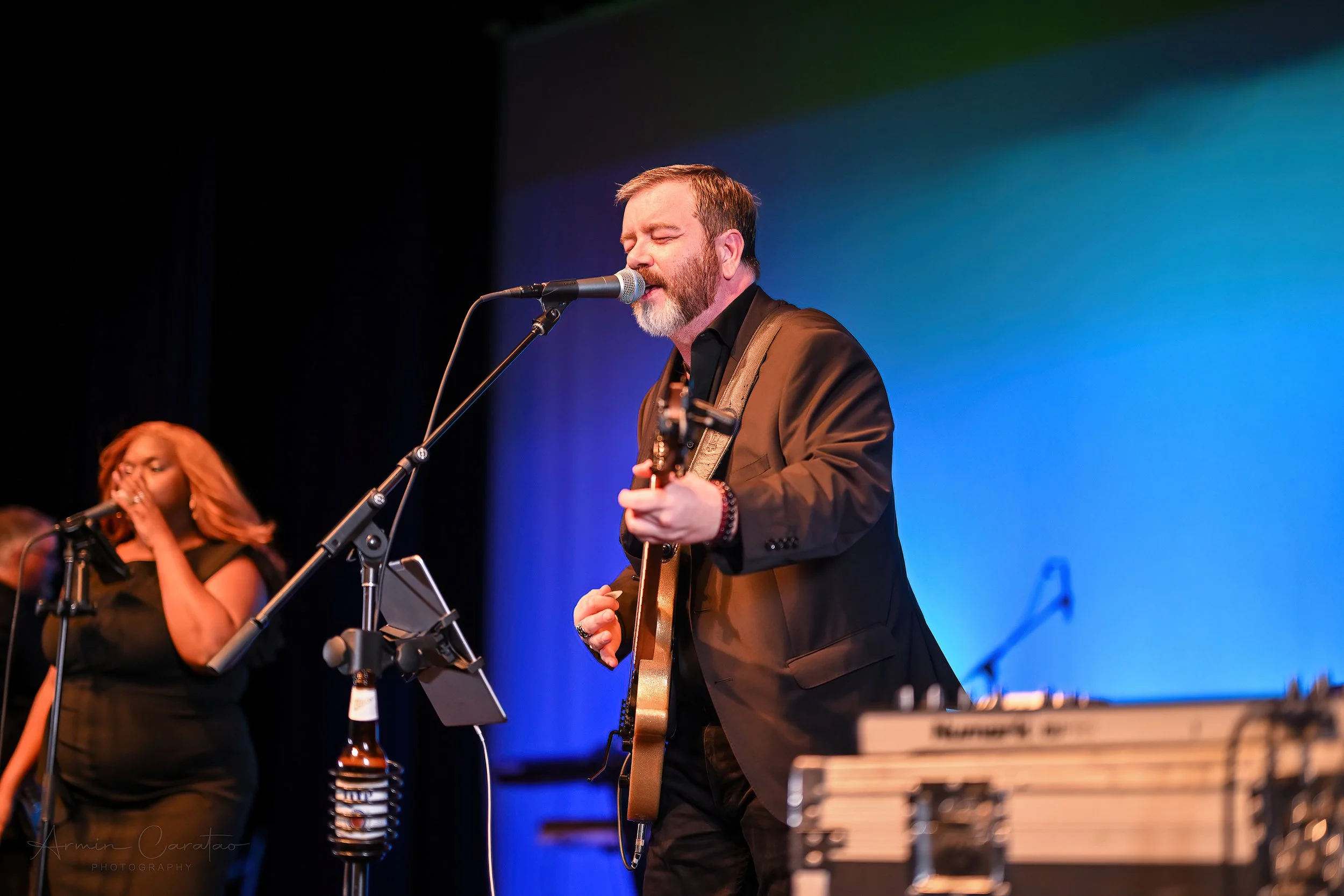 Male musician singing and playing guitar on stage, with a female backing vocalist and microphones visible, set against a blue-lit background.