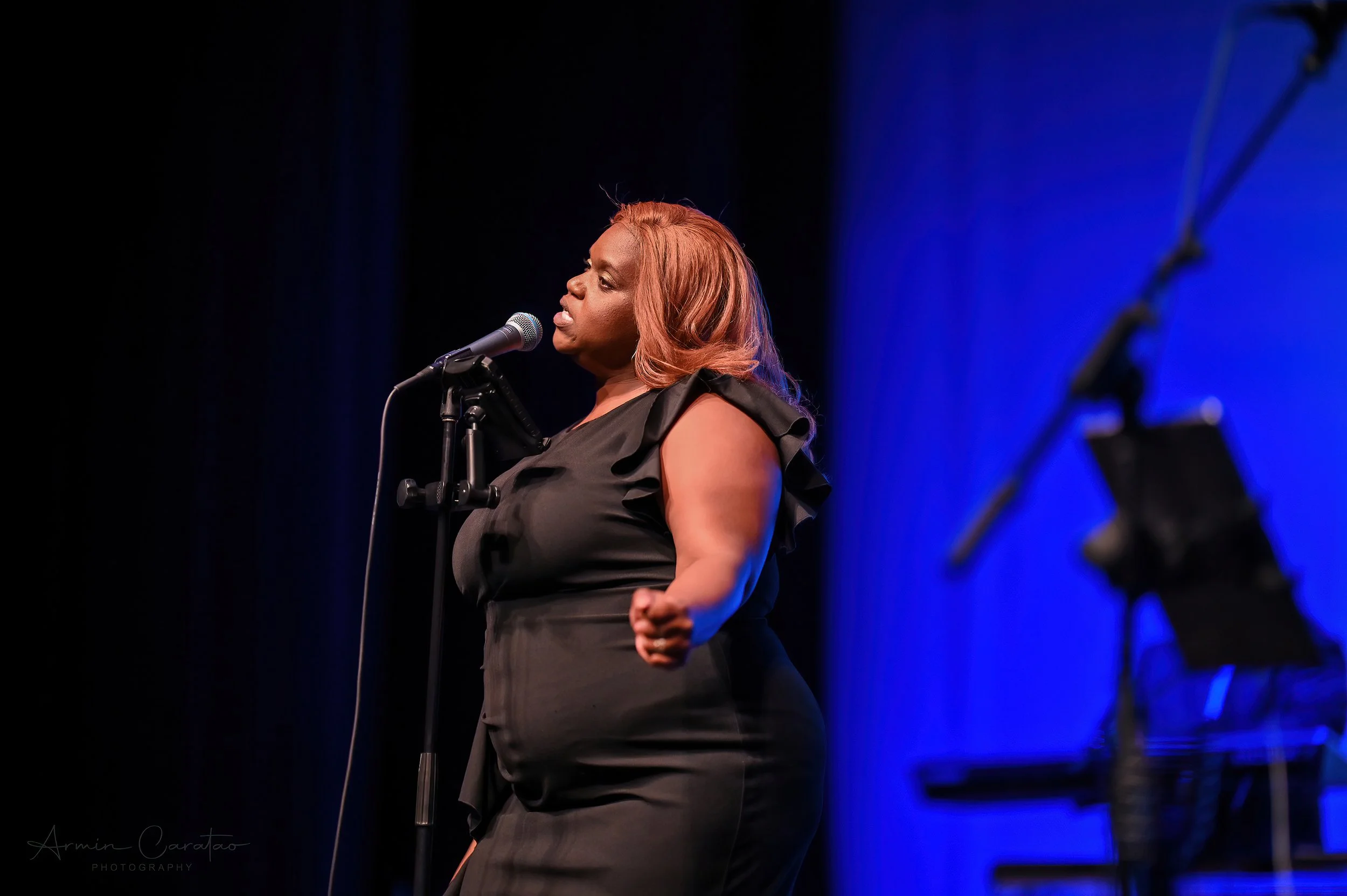 Woman with long hair singing into a microphone on stage with a blue background.