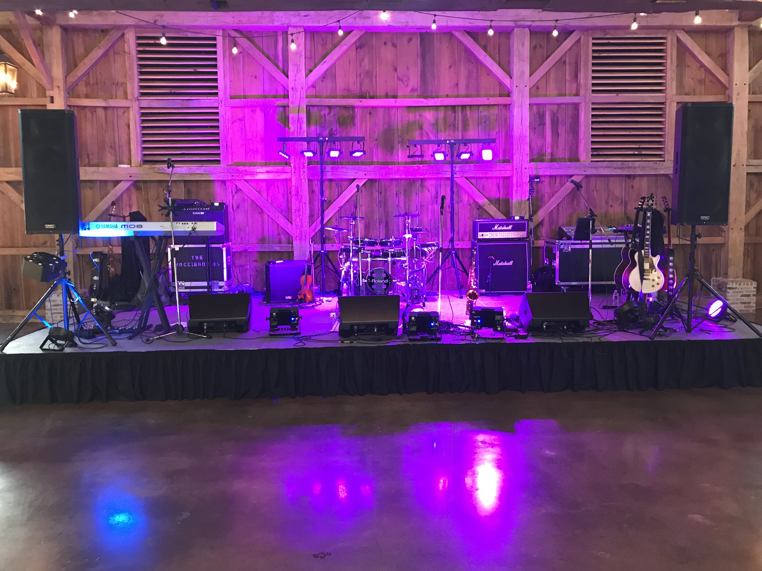 Stage setup with musical instruments and equipment, including a keyboard, guitars, drums, speakers, and amplifiers, with purple lighting in a wooden interior.