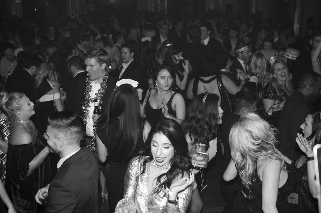 Black and white photo of a crowded party with people dancing and socializing, some wearing formal attire and festive decorations.