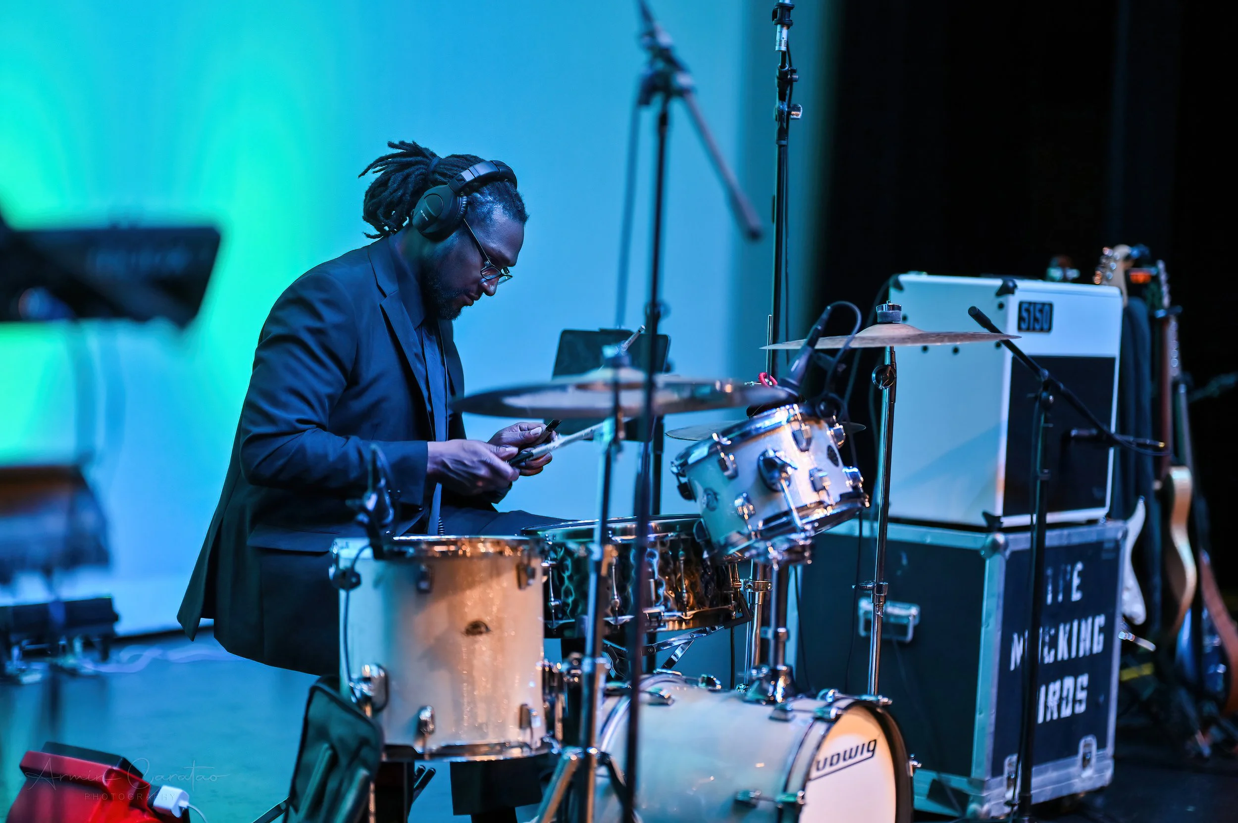 A musician wearing headphones tuning a drum set on stage with equipment in the background.