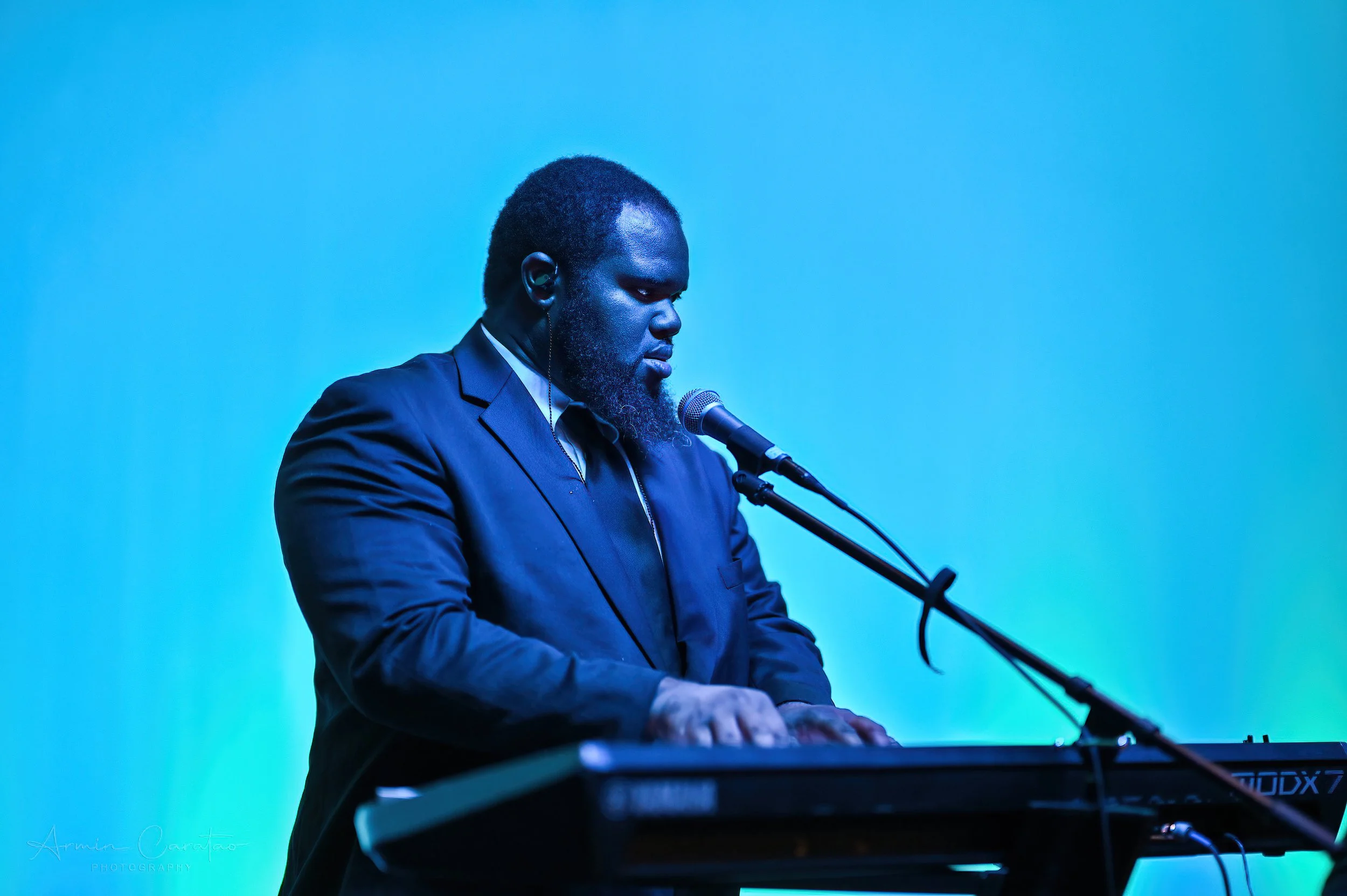 Man in a suit playing a keyboard and singing into a microphone against a blue background.