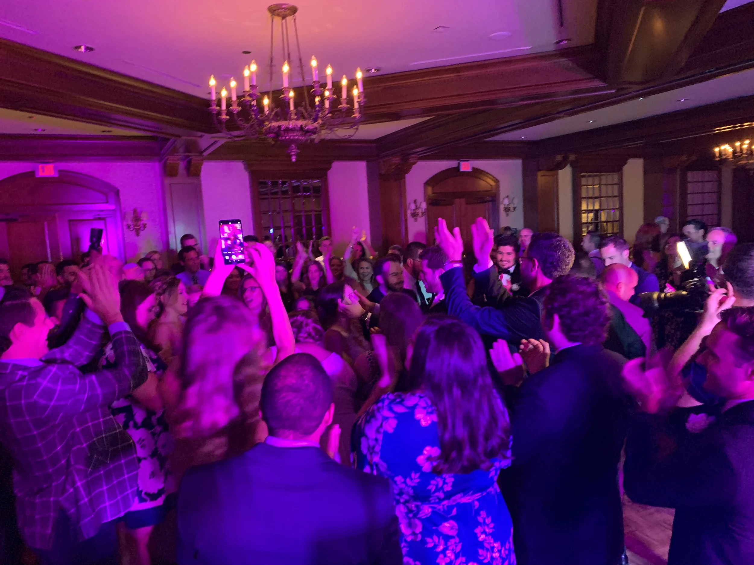 A large group of people dancing and celebrating at an indoor party under a chandelier, with a raised hand capturing a photo on a smartphone.