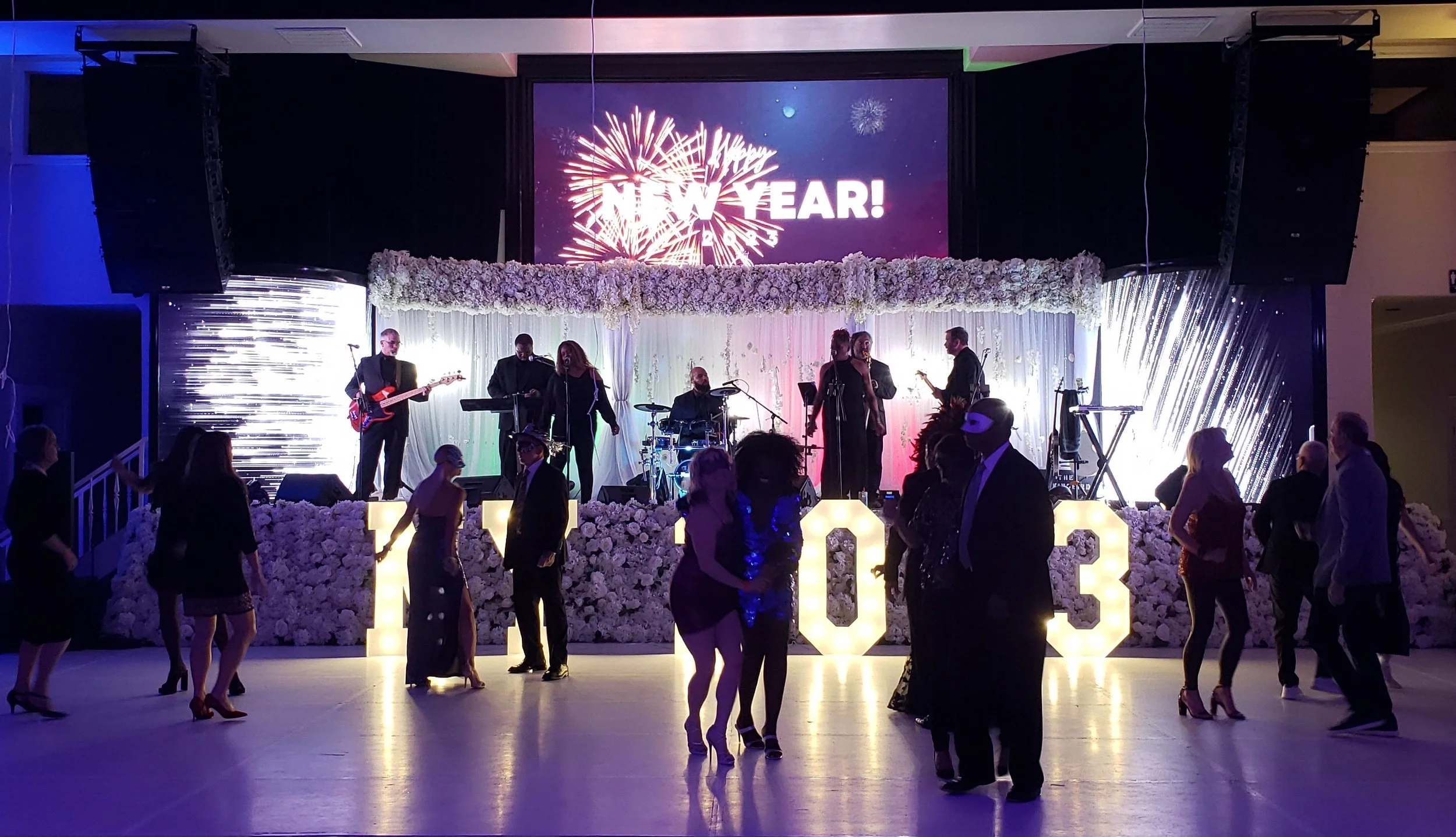 People celebrating at a New Year's event with a live band on stage, dancers on the floor, and a "NEW YEAR!" fireworks display projected. The stage is decorated with flowers and illuminated numbers "2023."