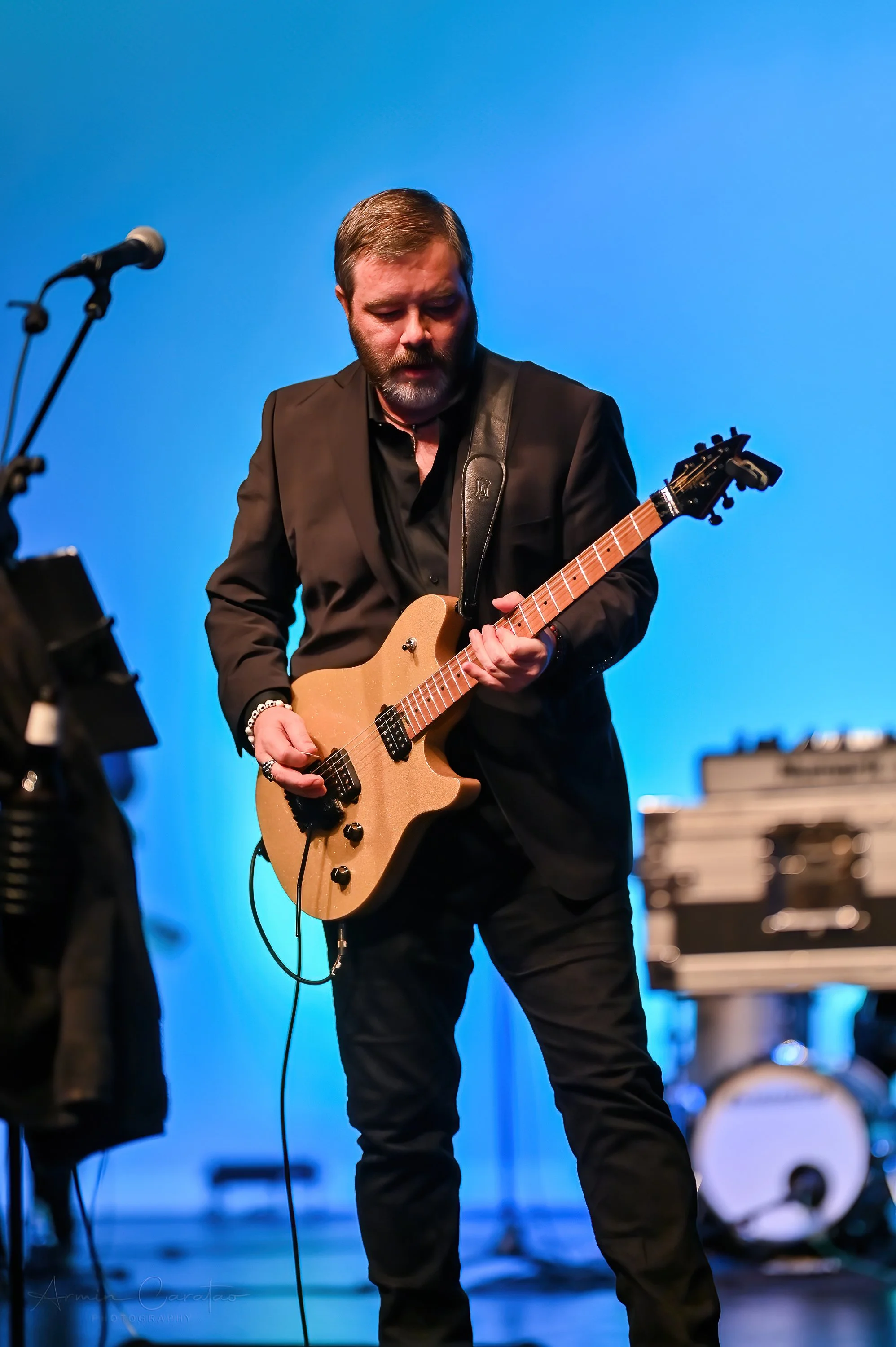 A man in a black suit playing an electric guitar on stage with a blue background.
