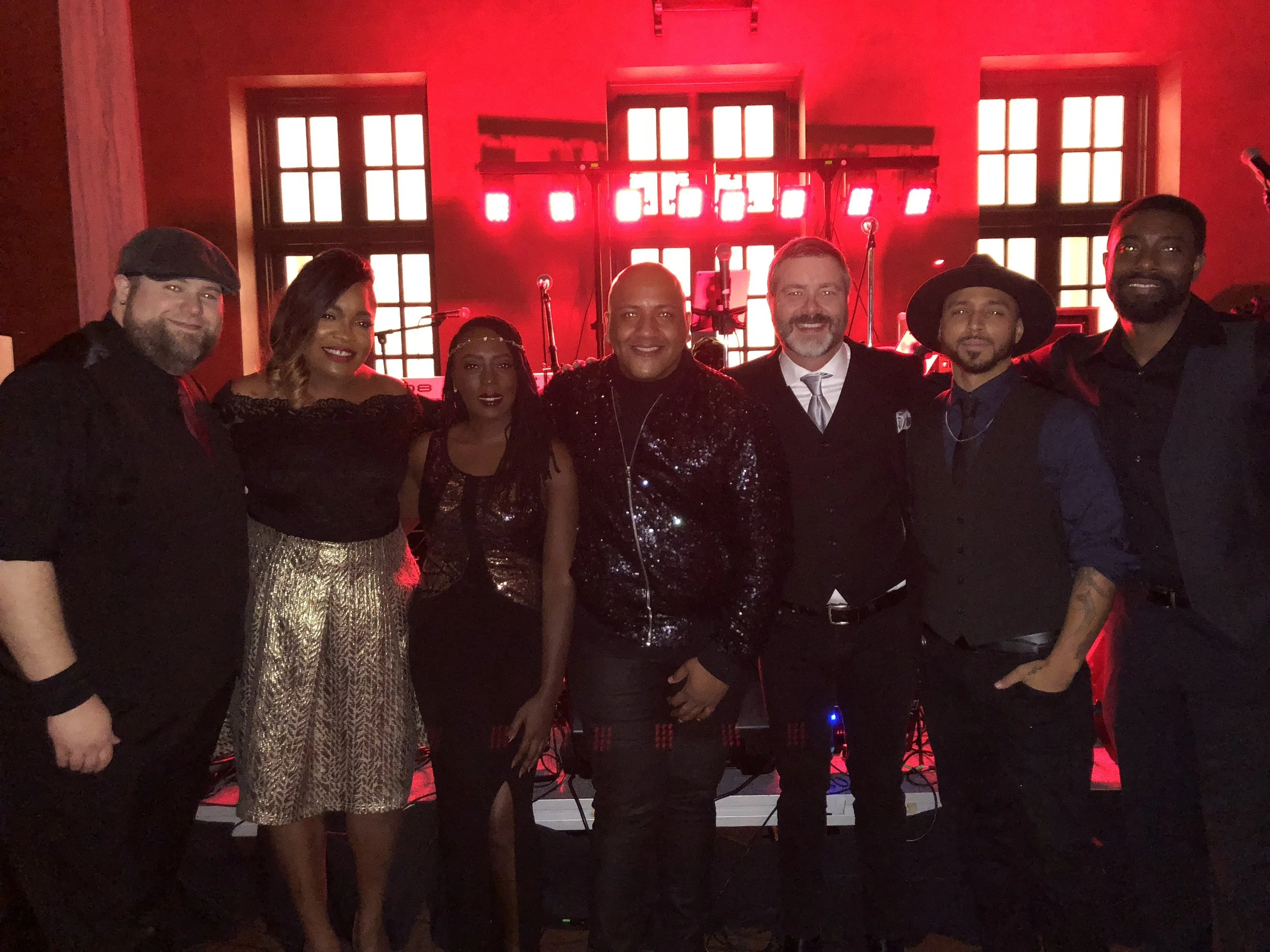 Group photo of seven people dressed in formal and semi-formal attire standing in front of a stage with red lighting.