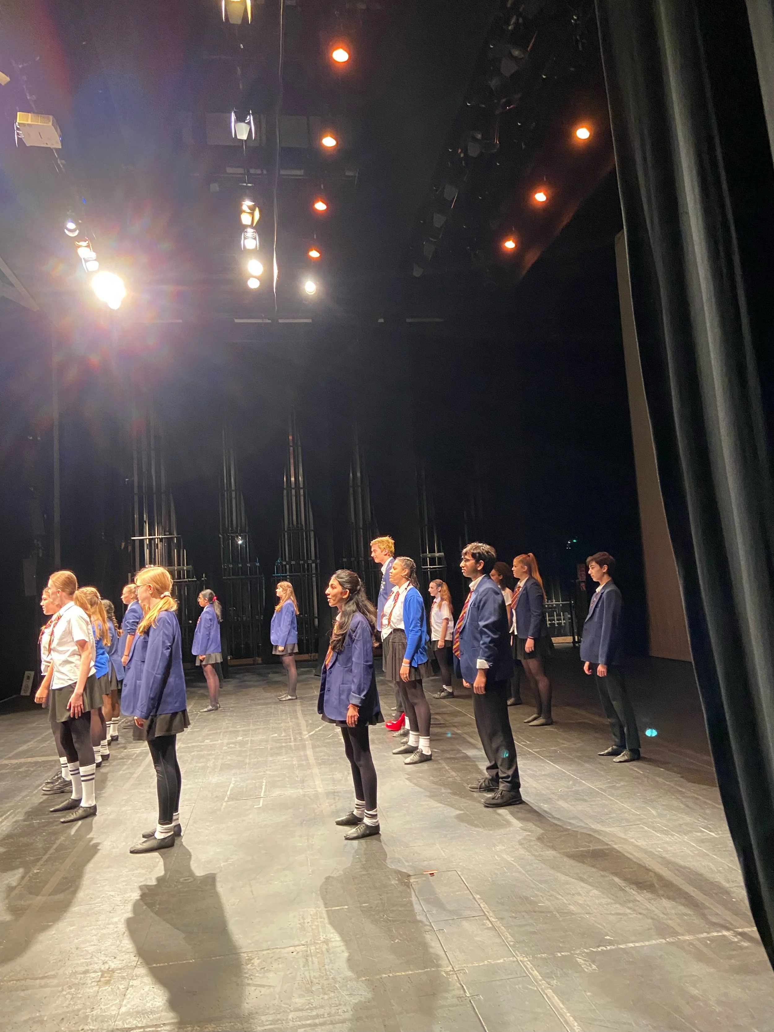 Students standing on stage in school uniforms during rehearsal or performance.