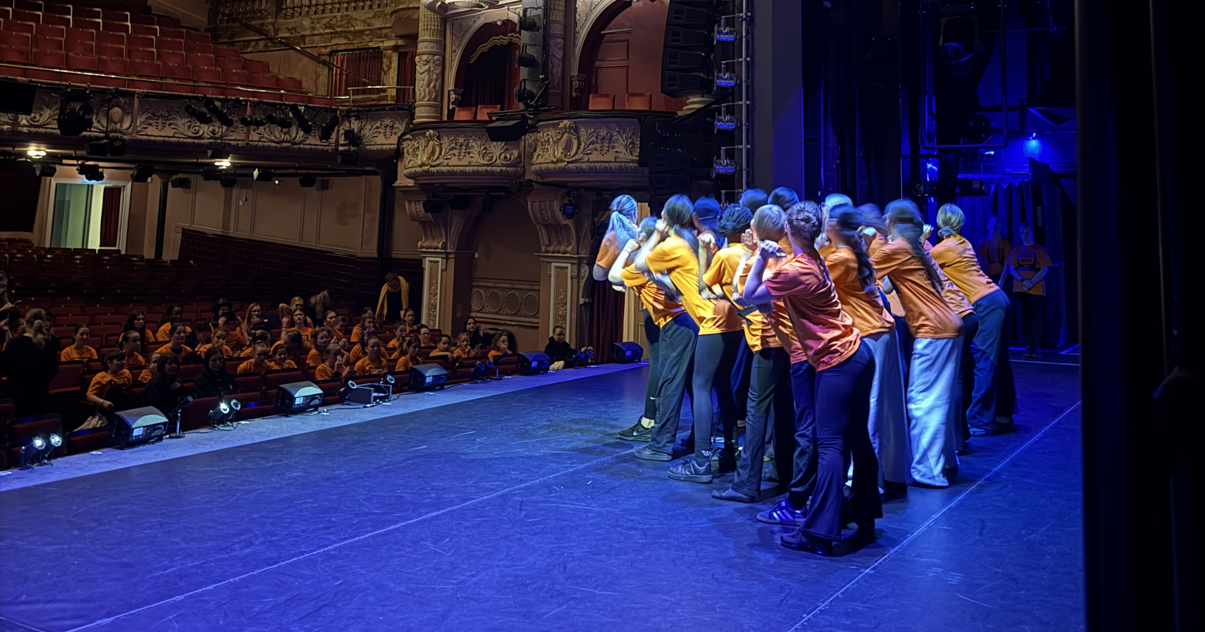 A group of performers dressed in orange shirts on stage bowing to an audience in a theater. The audience, also mostly dressed in orange, is sitting in red seats watching the performance.