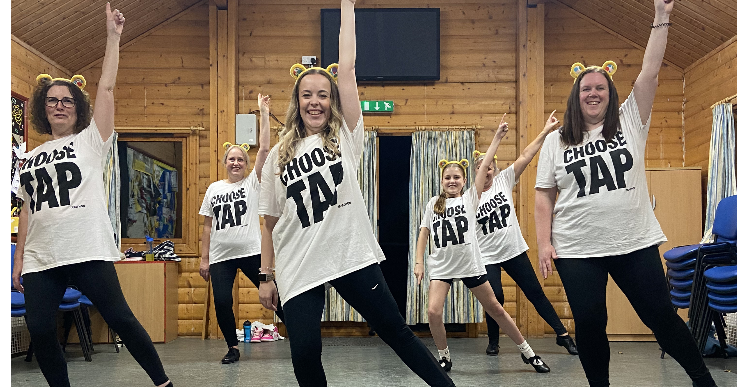 A group of women wearing matching white T-shirts with the words 'CHOOSE TAP' and black pants, dancing in a room with wooden walls. Some women are wearing yellow bear ear headbands, and they are smiling with arms raised.