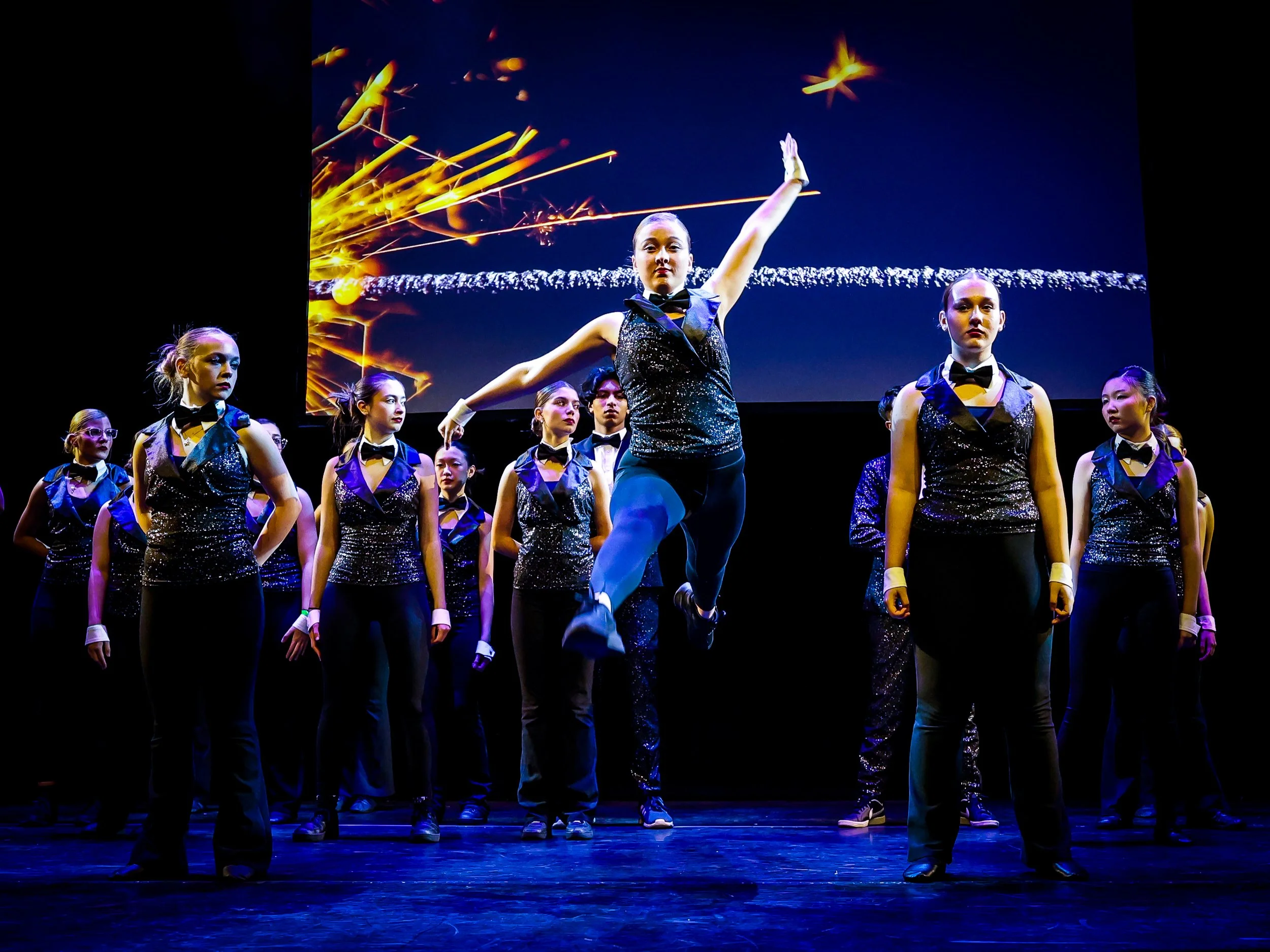 A group of young female dancers, dressed in black and sparkly costumes with bow ties, perform on stage with a digital backdrop of fireworks.