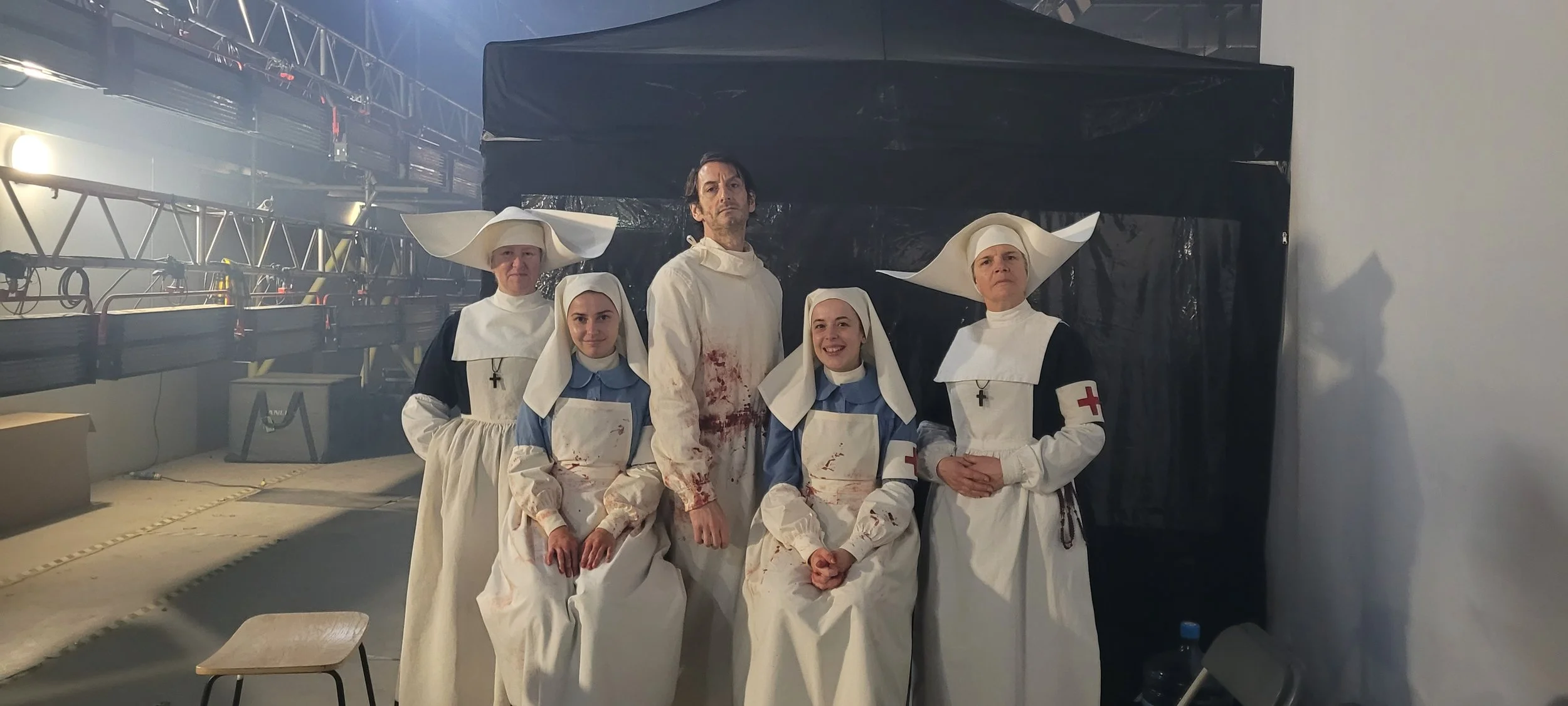 People dressed as nurses and a man in a blood-stained coat and apron, standing in front of a dark backdrop, on a set or staging area.