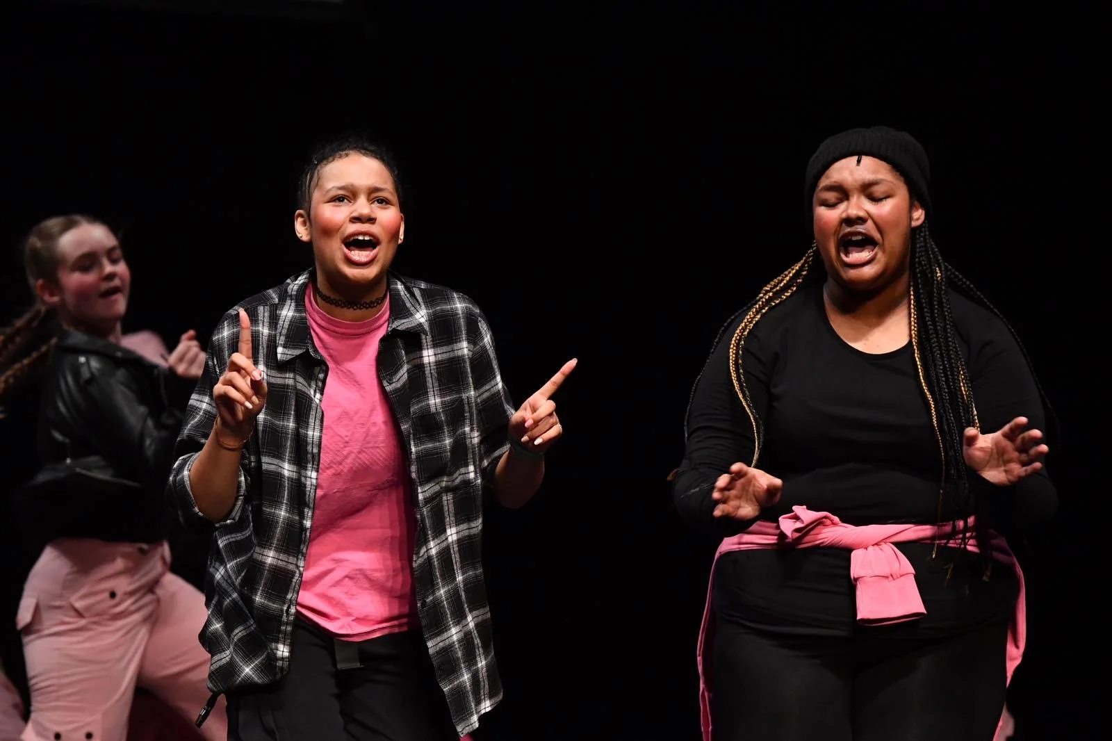 Three young women performing on stage, singing passionately, with a dark background; two are wearing black tops with pink accents, and one is in a pink shirt with a plaid jacket.
