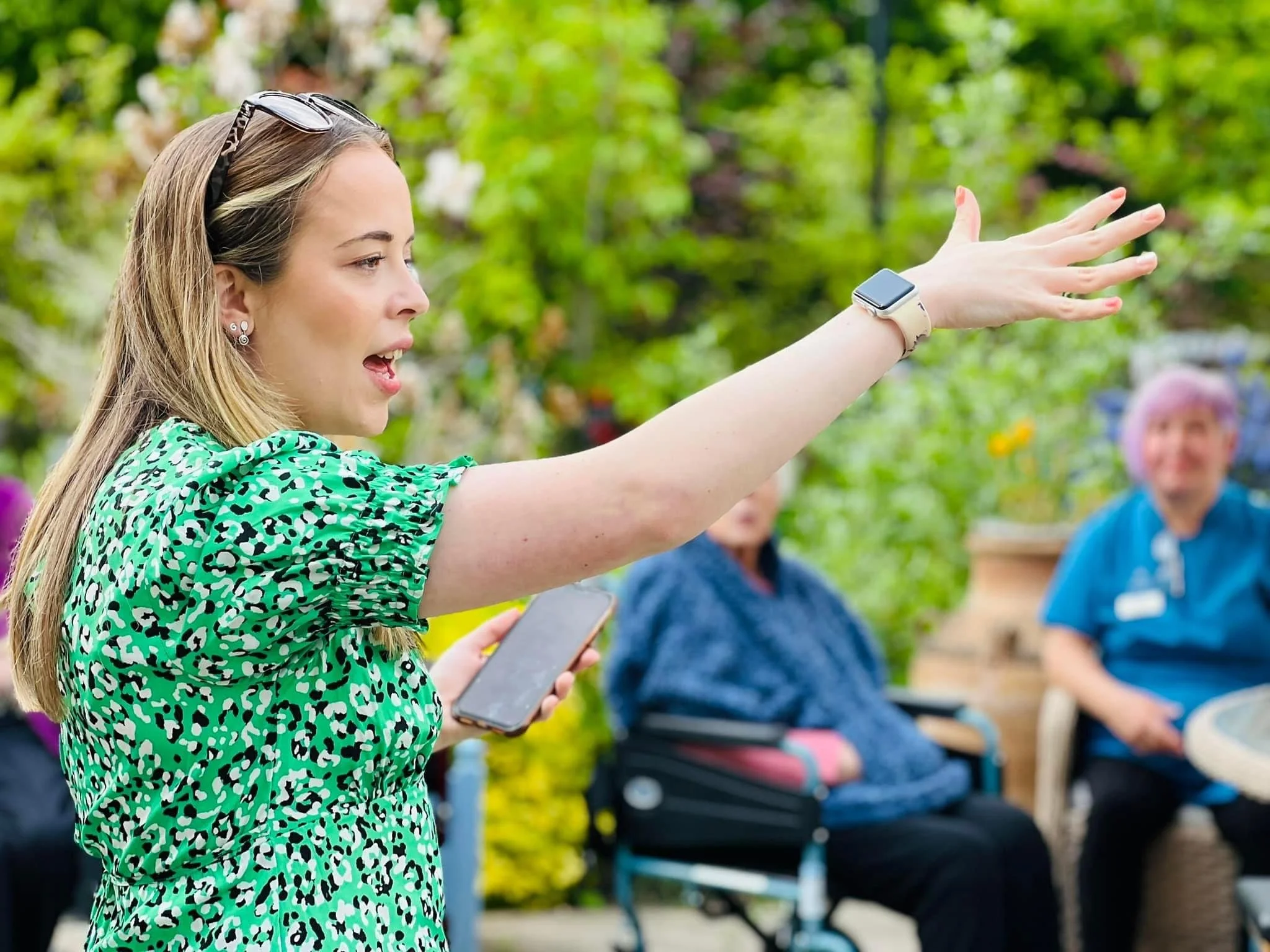 A woman with long blonde hair, wearing a green patterned dress and a watch, is speaking and gesturing with her right arm extended outward. She holds a smartphone in her left hand. In the background, there are other women sitting outdoors in a garden 